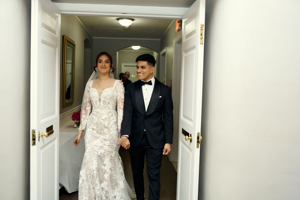 A couple walks down a hallway, the bride wearing a long-sleeved lace gown and the groom in a classic black tuxedo at 3 west Club