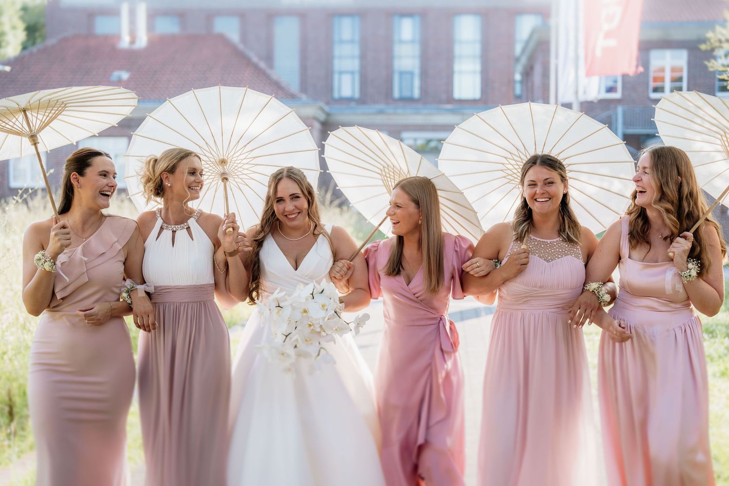 Bridal party in pink dresses poses with white parasols against a brick building backdrop.