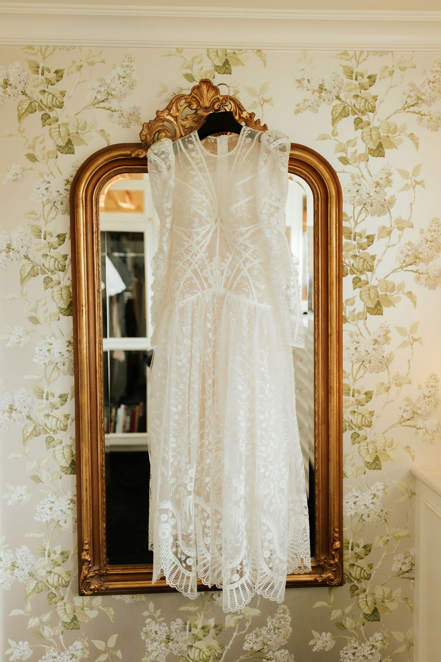 White lace dress hanging against floral wallpaper next to ornate gold-framed mirrors creates a vintage romantic atmosphere.