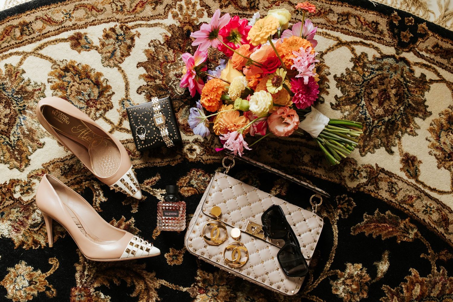 Wedding details including pink and orange flowers, designer shoes, and perfume bottles arranged on an ornate patterned rug.
