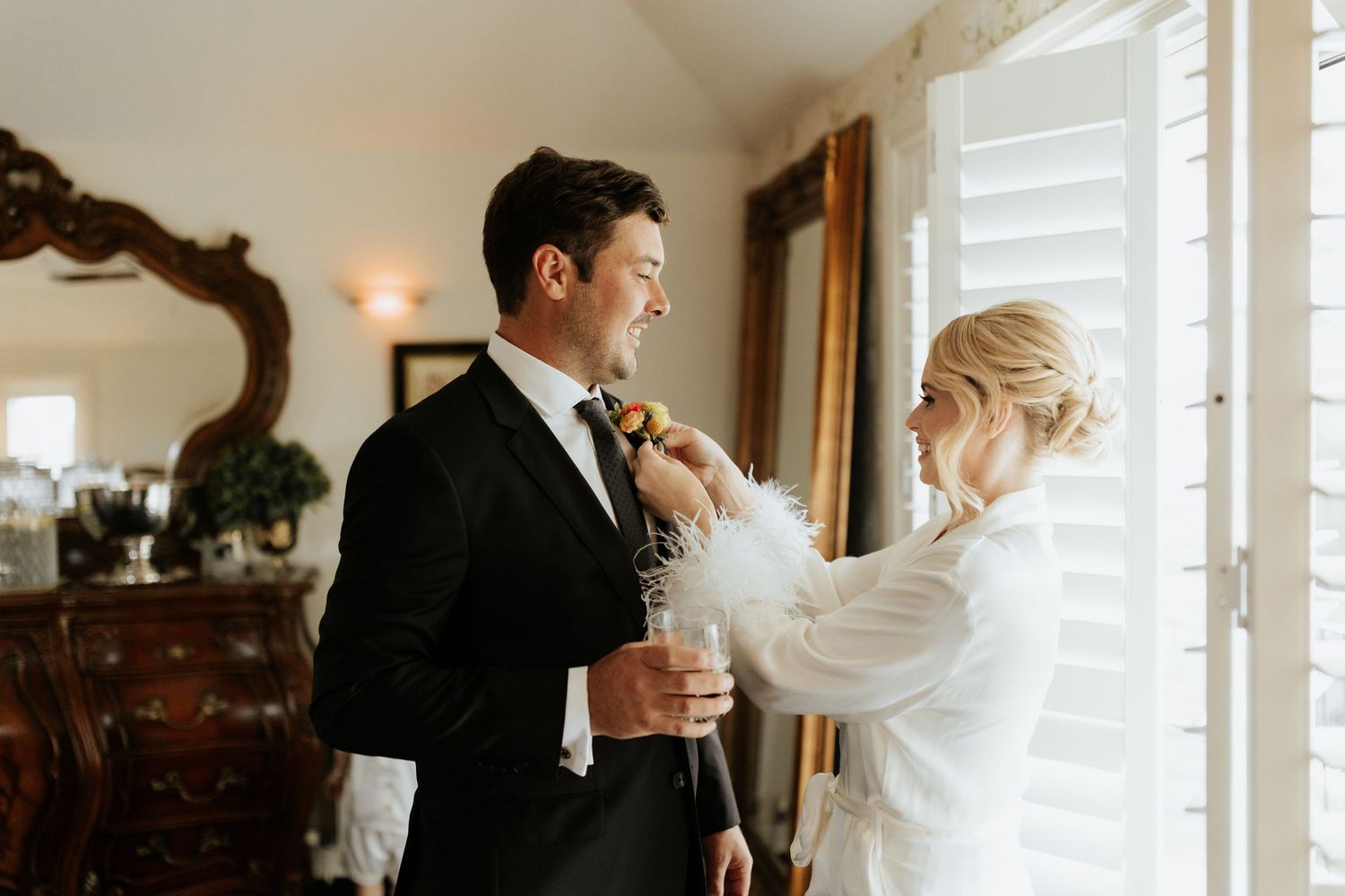 A couple shares a tender moment while getting ready on their wedding day in an elegant room with vintage furniture.