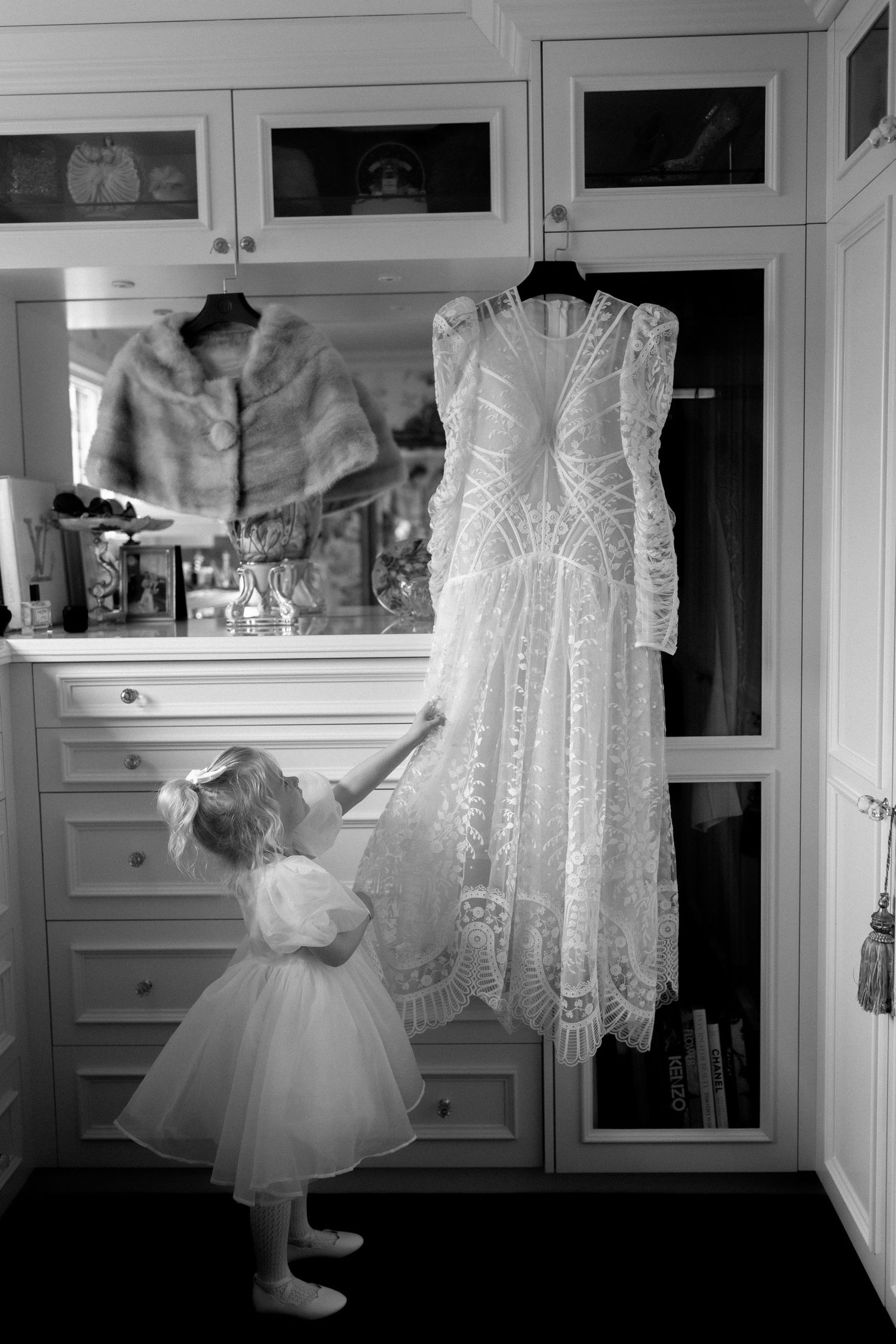 Black and white photo of a wedding dress hanging while a small child in tulle dress reaches up towards it.