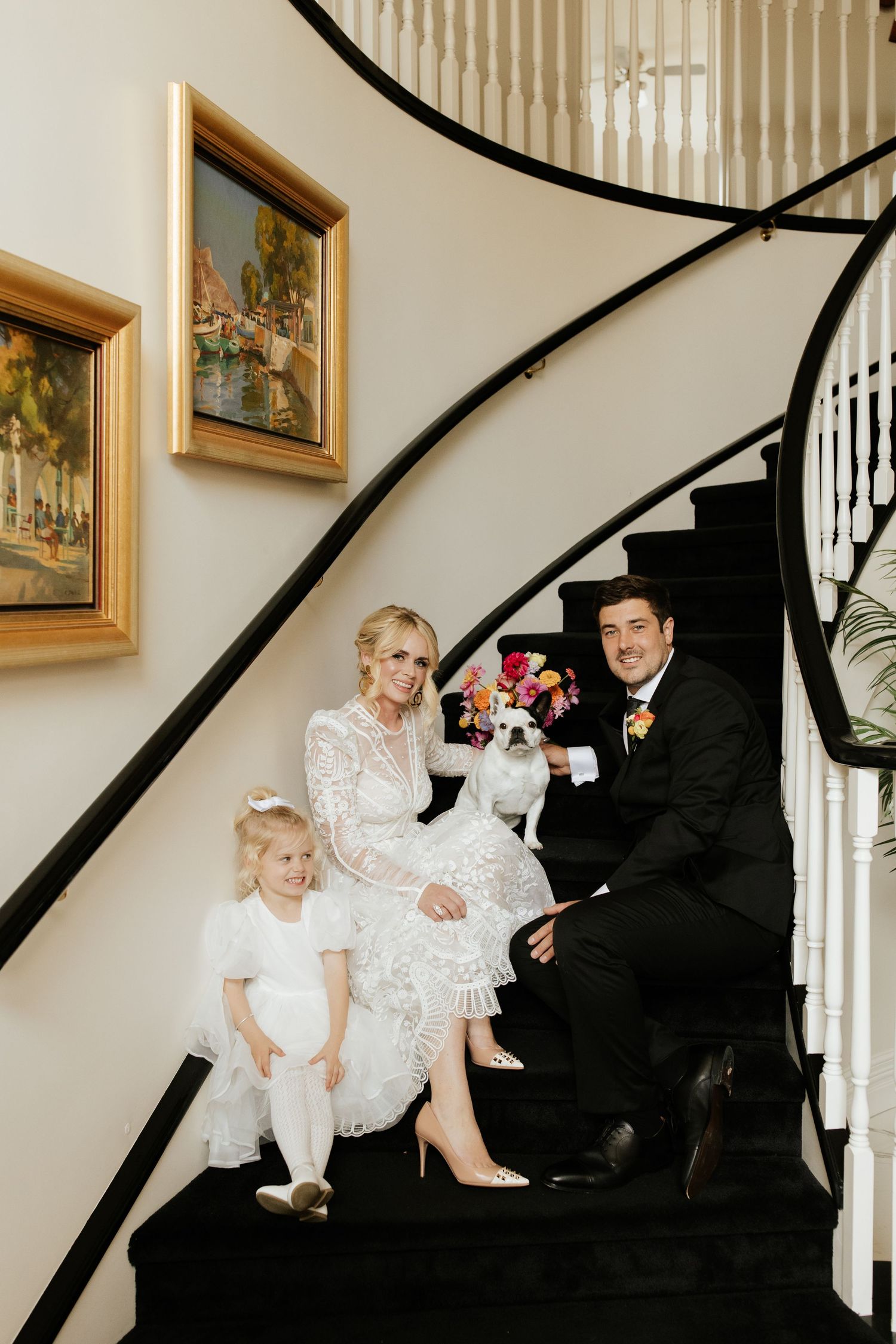 A wedding portrait on elegant black stairs featuring a couple in formal attire with a small child and dog.
