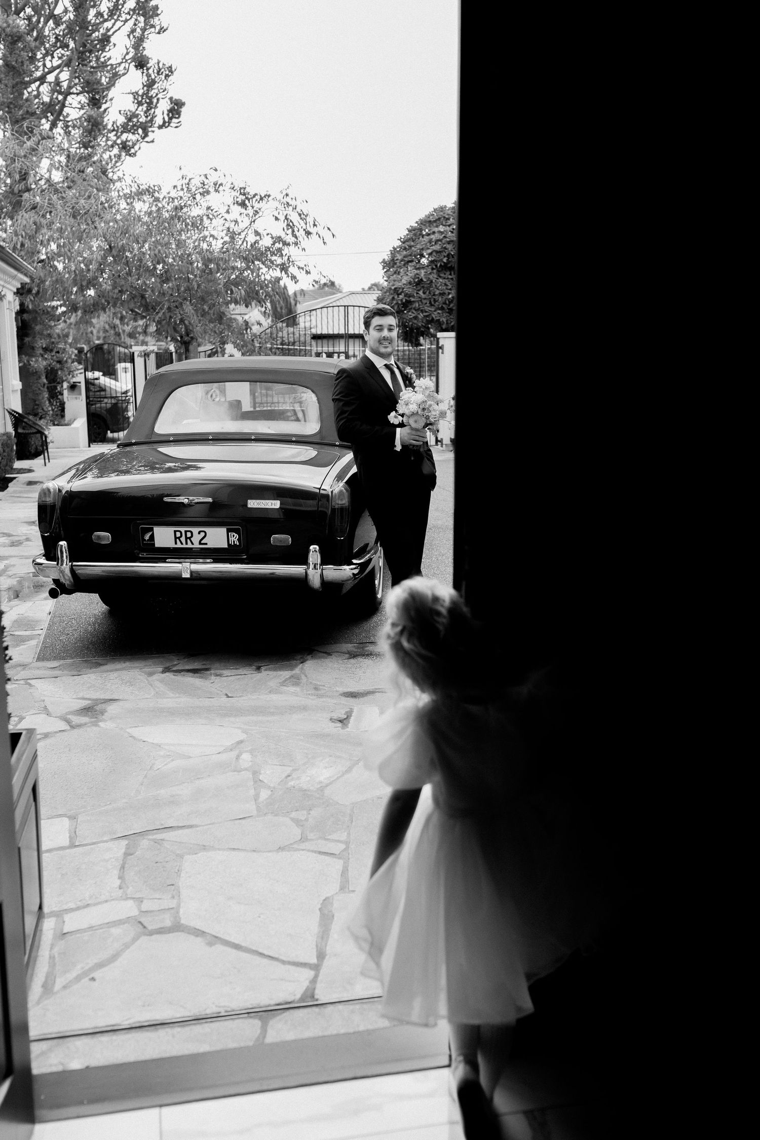 A black and white wedding photo with a vintage car and formal attire in a driveway.