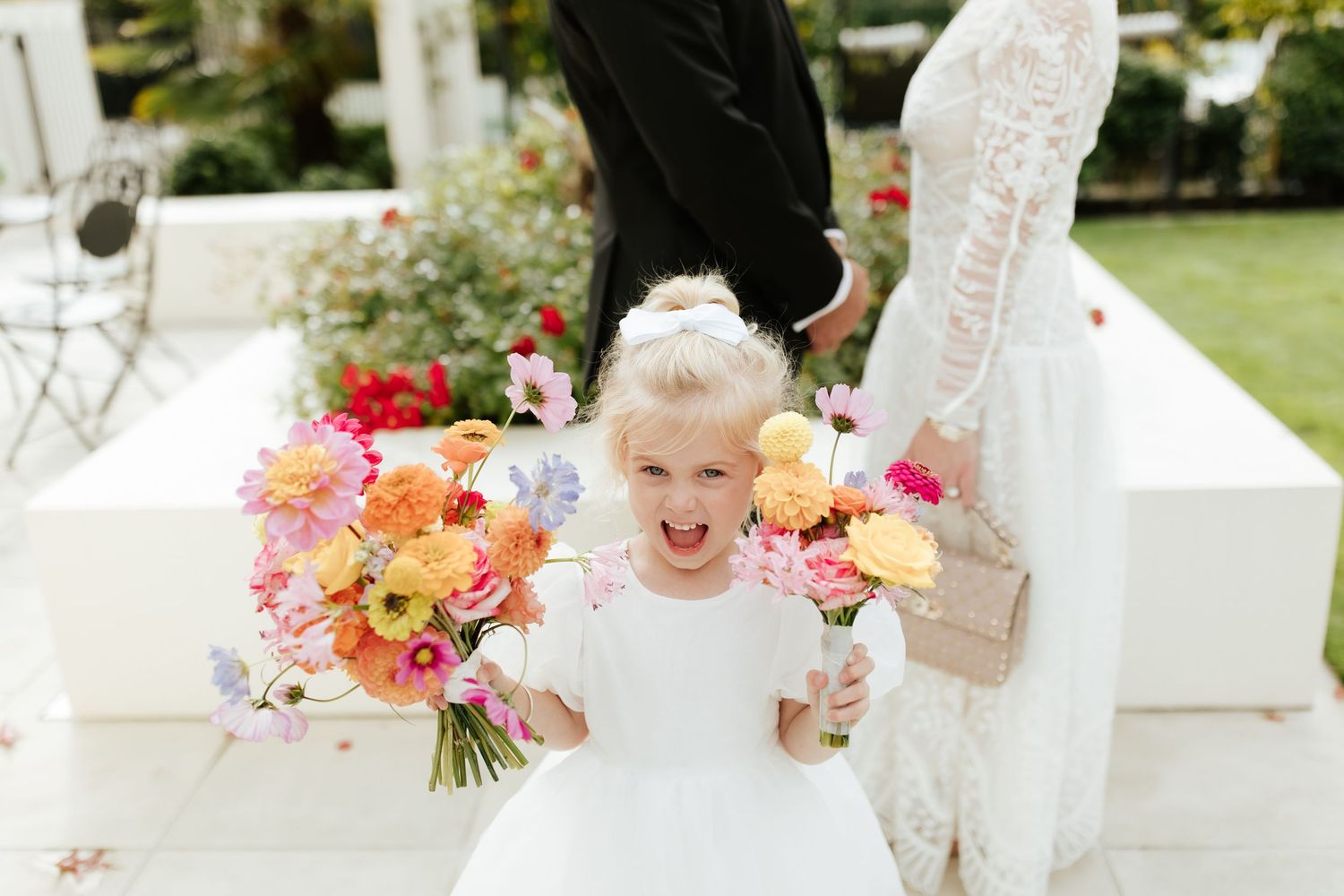 An excited child in a white dress stands between two large colorful floral bouquets at an outdoor wedding.