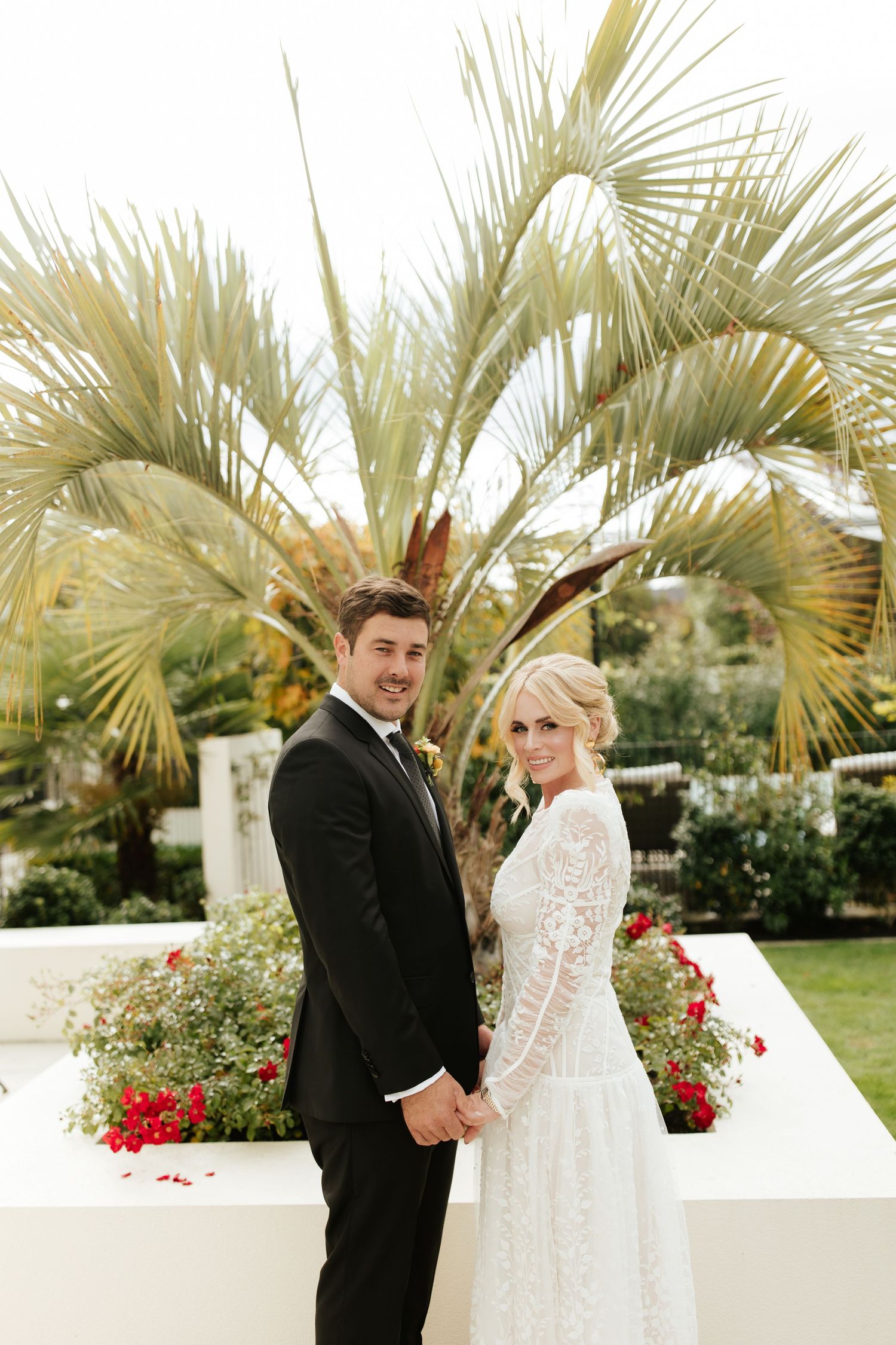 A couple pose together in elegant wedding attire in front of palm trees and floral landscaping.