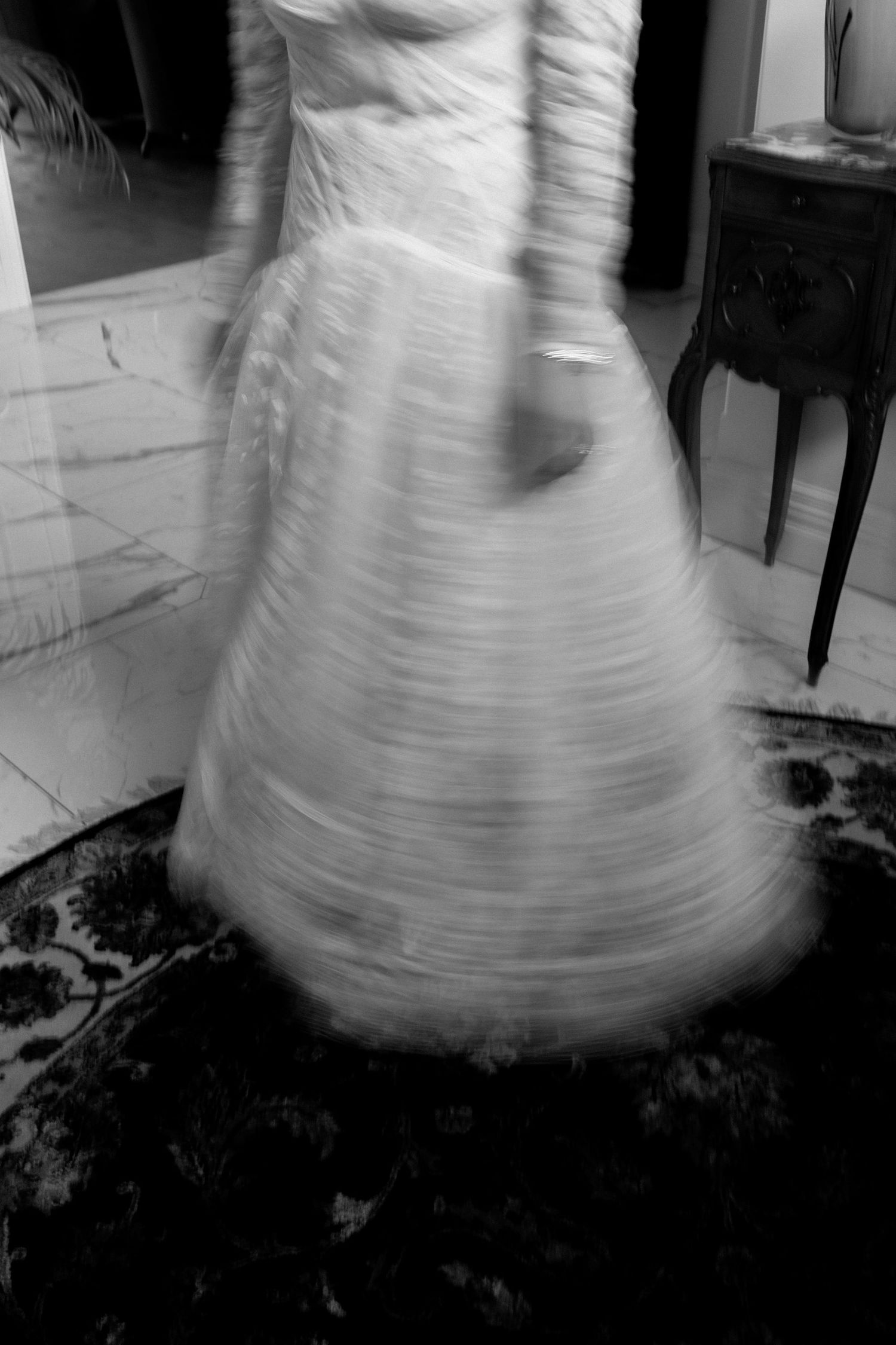 A black and white motion-blurred photograph of a spinning wedding dress on a dark patterned rug.