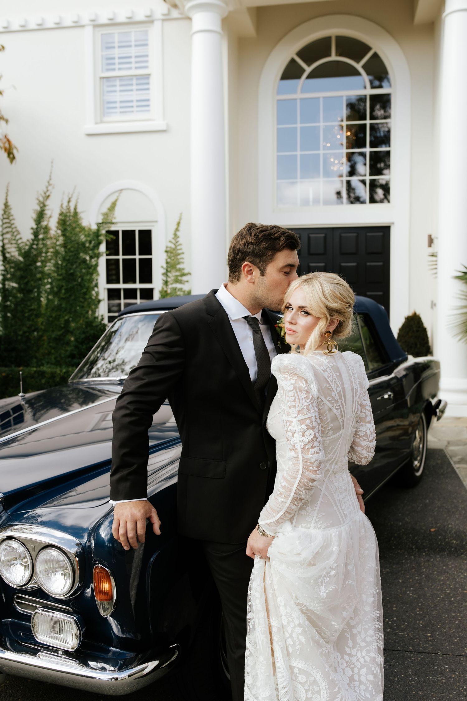 Wedding couple poses beside classic vintage car in front of white colonial building.