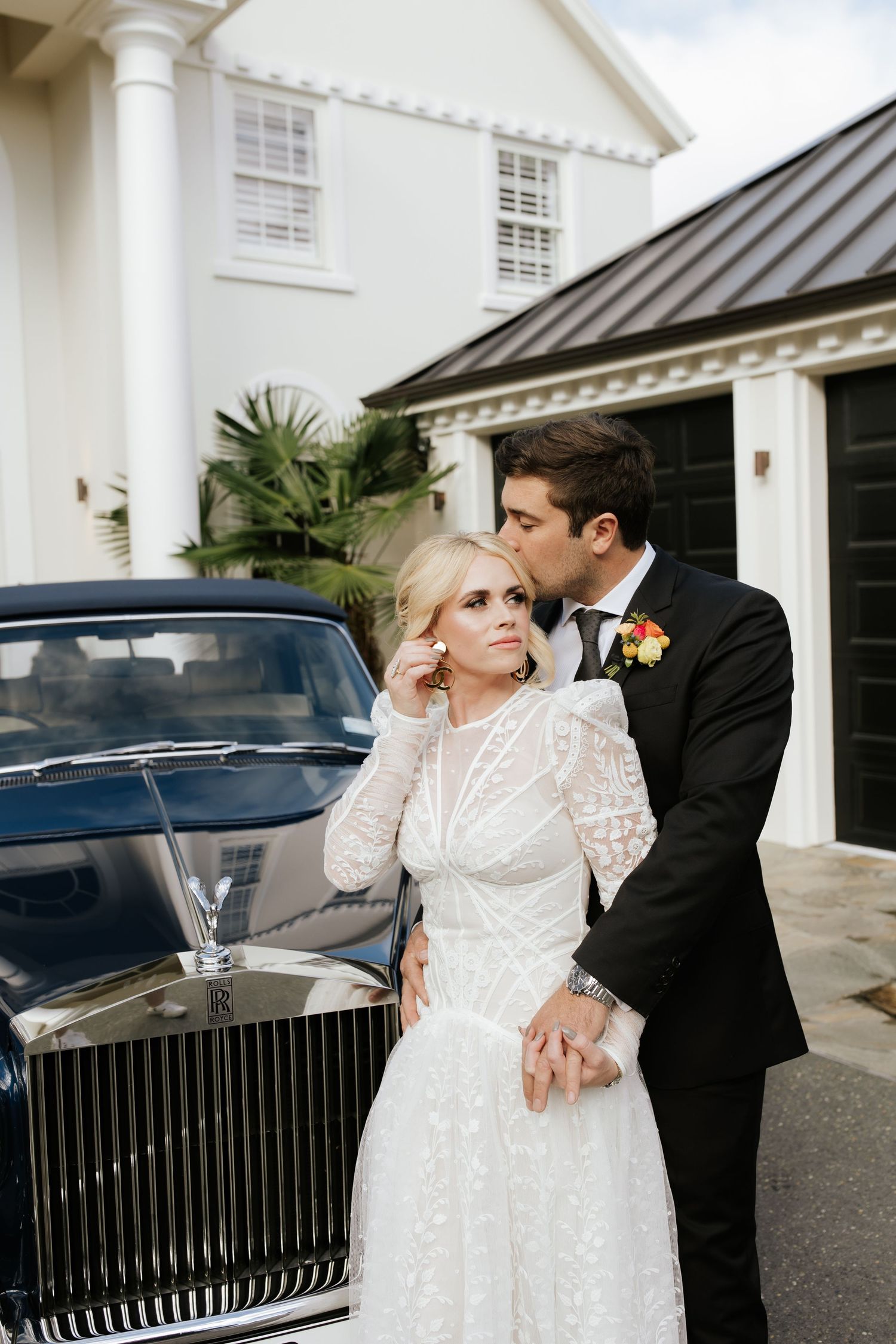 A romantic wedding couple poses next to a vintage Rolls Royce car in front of a white colonial style building.