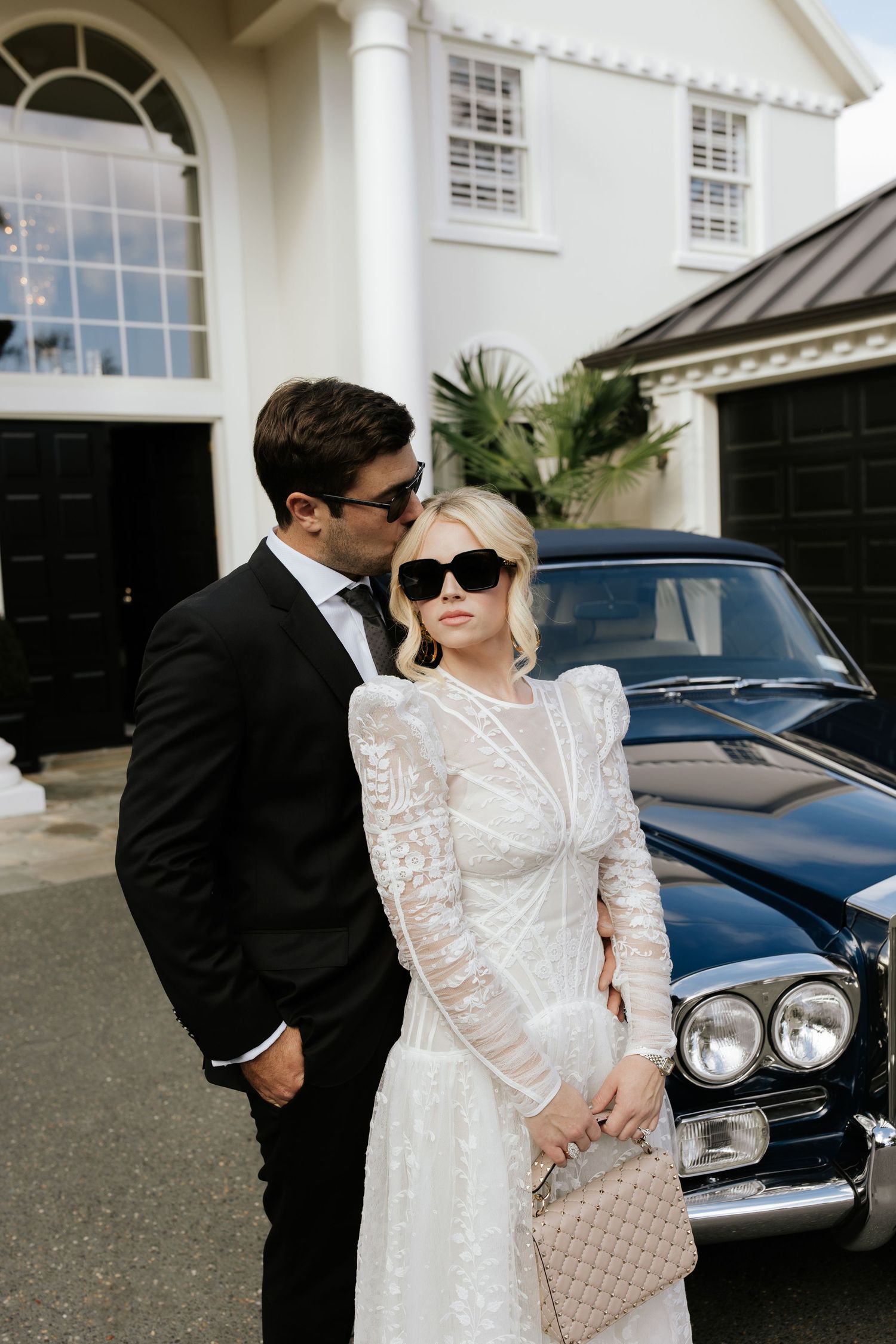 Elegant wedding couple posing next to vintage car outside white building with architectural details.