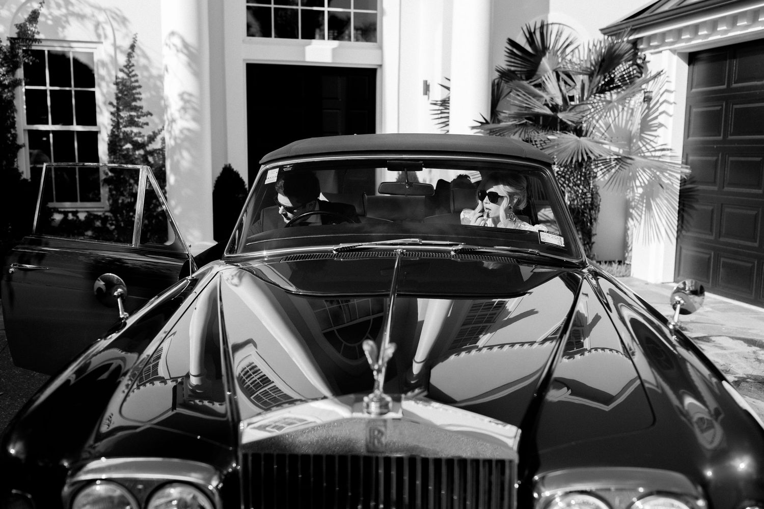 Vintage Rolls-Royce with ornate hood ornament parked in front of elegant white mansion with tall windows.
