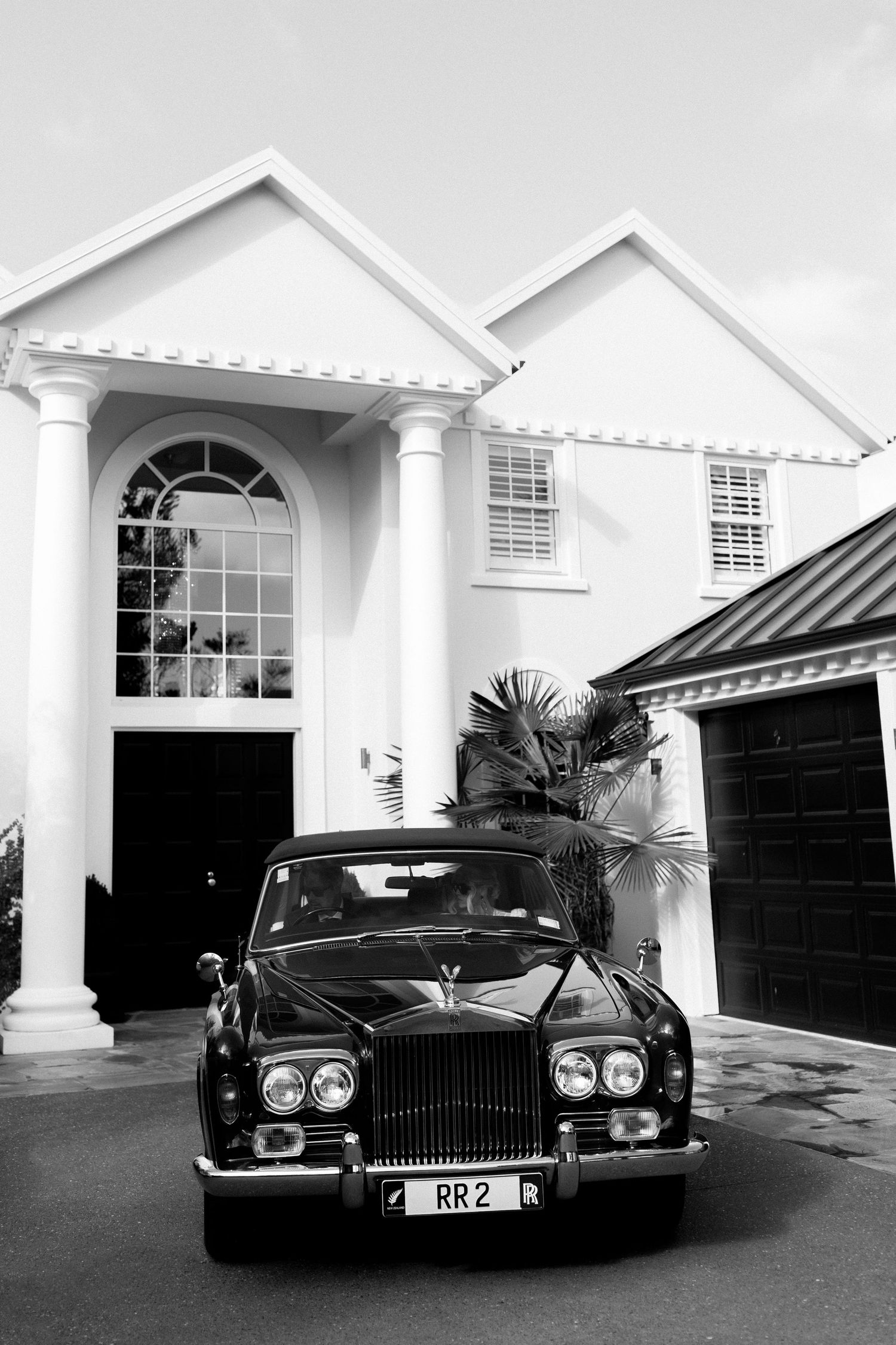 Classic black Rolls-Royce parked in front of white colonial style house with arched entrance and columns.