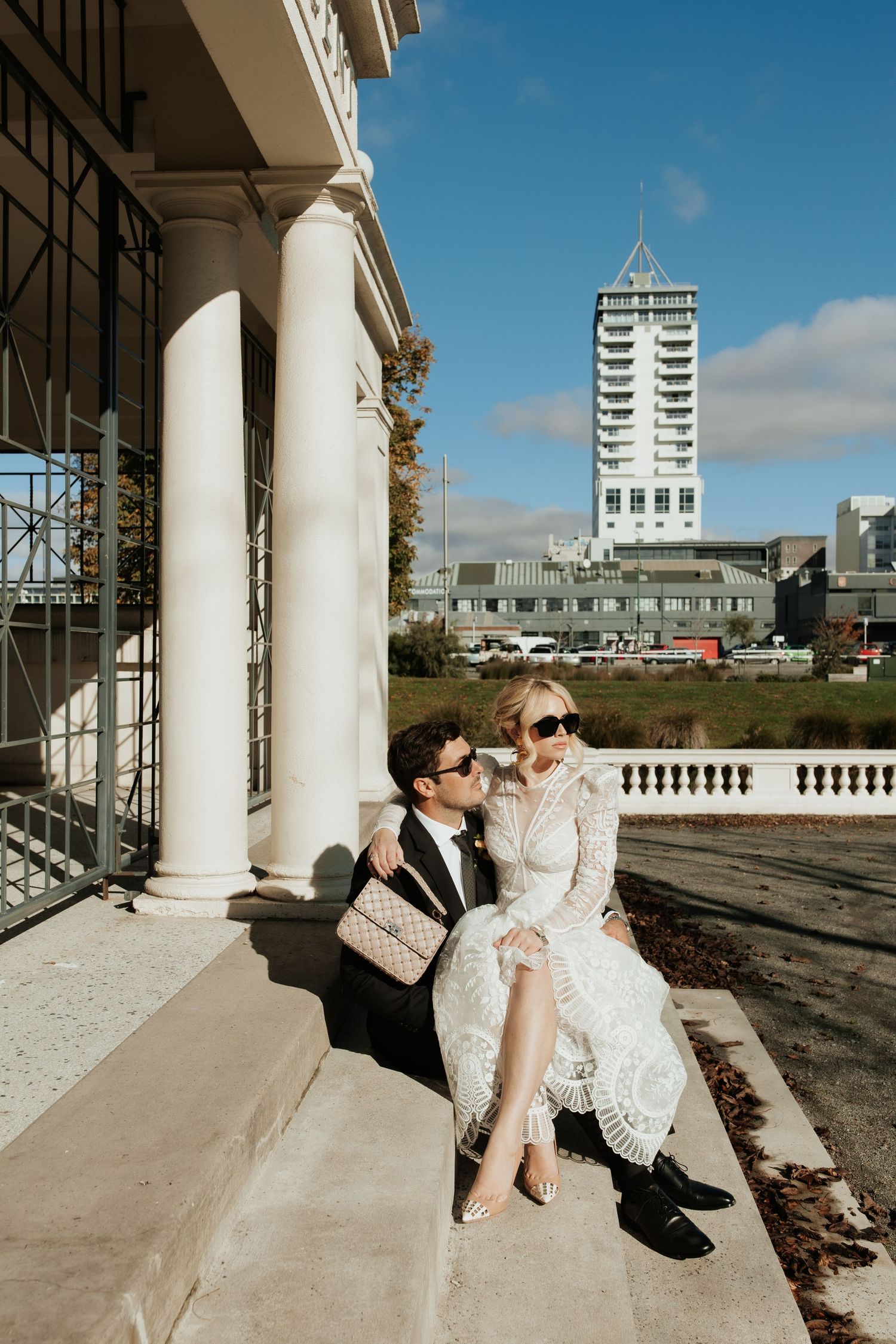 A couple in formal attire sits on steps with white columns and a tall tower building visible in the background.