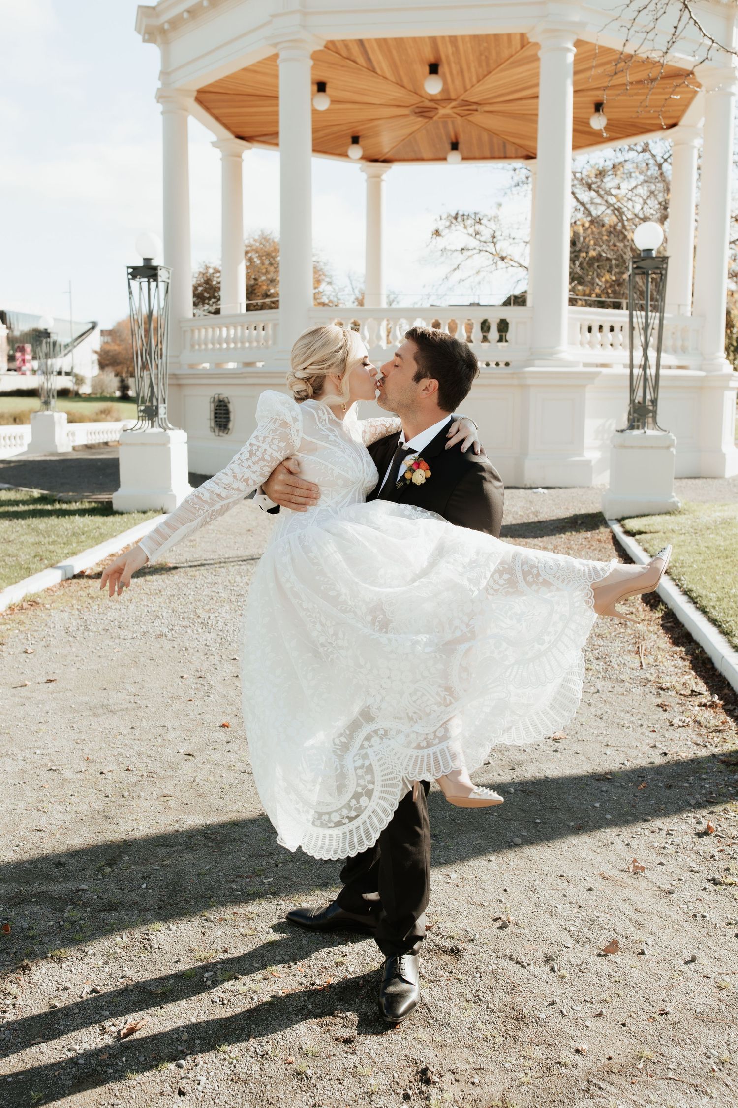 Joyful wedding moment captured at white gazebo as groom lifts bride in lace dress.