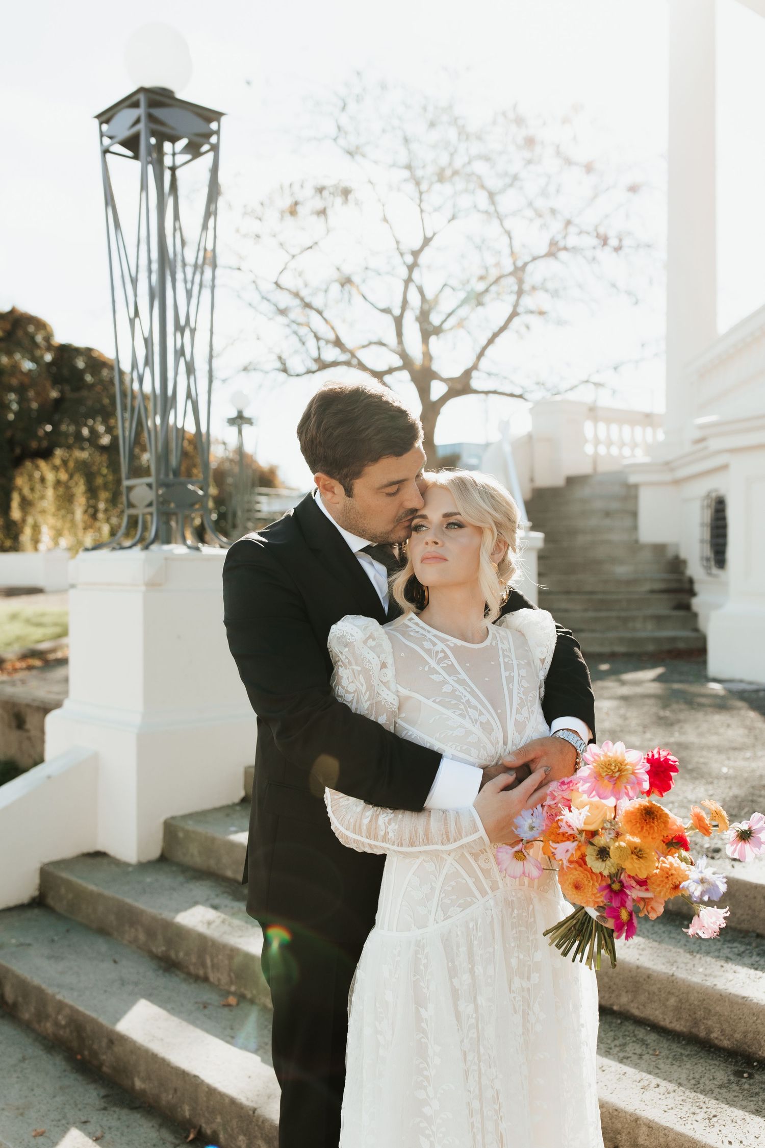 Romantic wedding couple embracing on white stone steps with colorful autumn bouquet.
