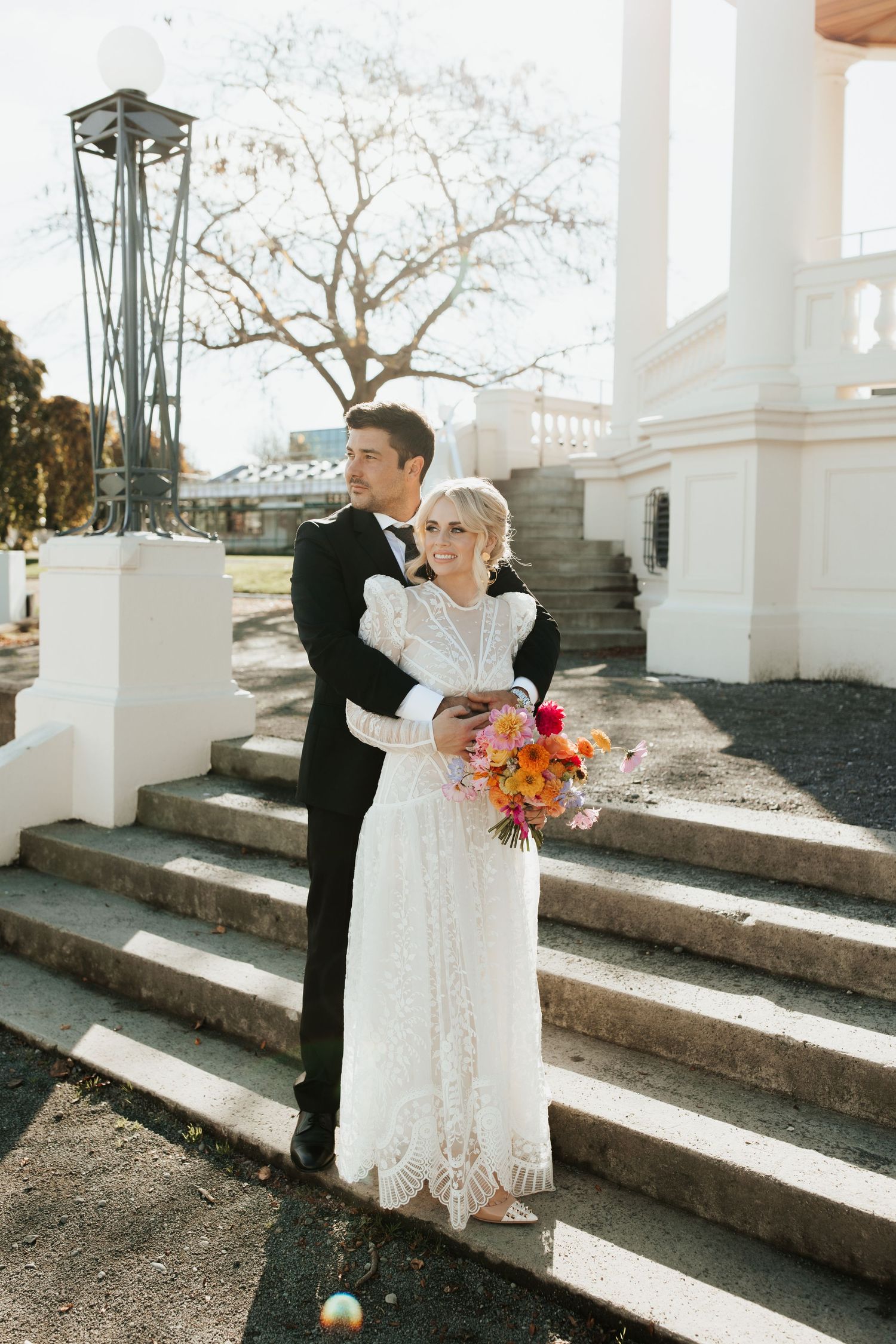 A couple stands together on stone steps while holding a vibrant fall-colored bouquet on their wedding day.