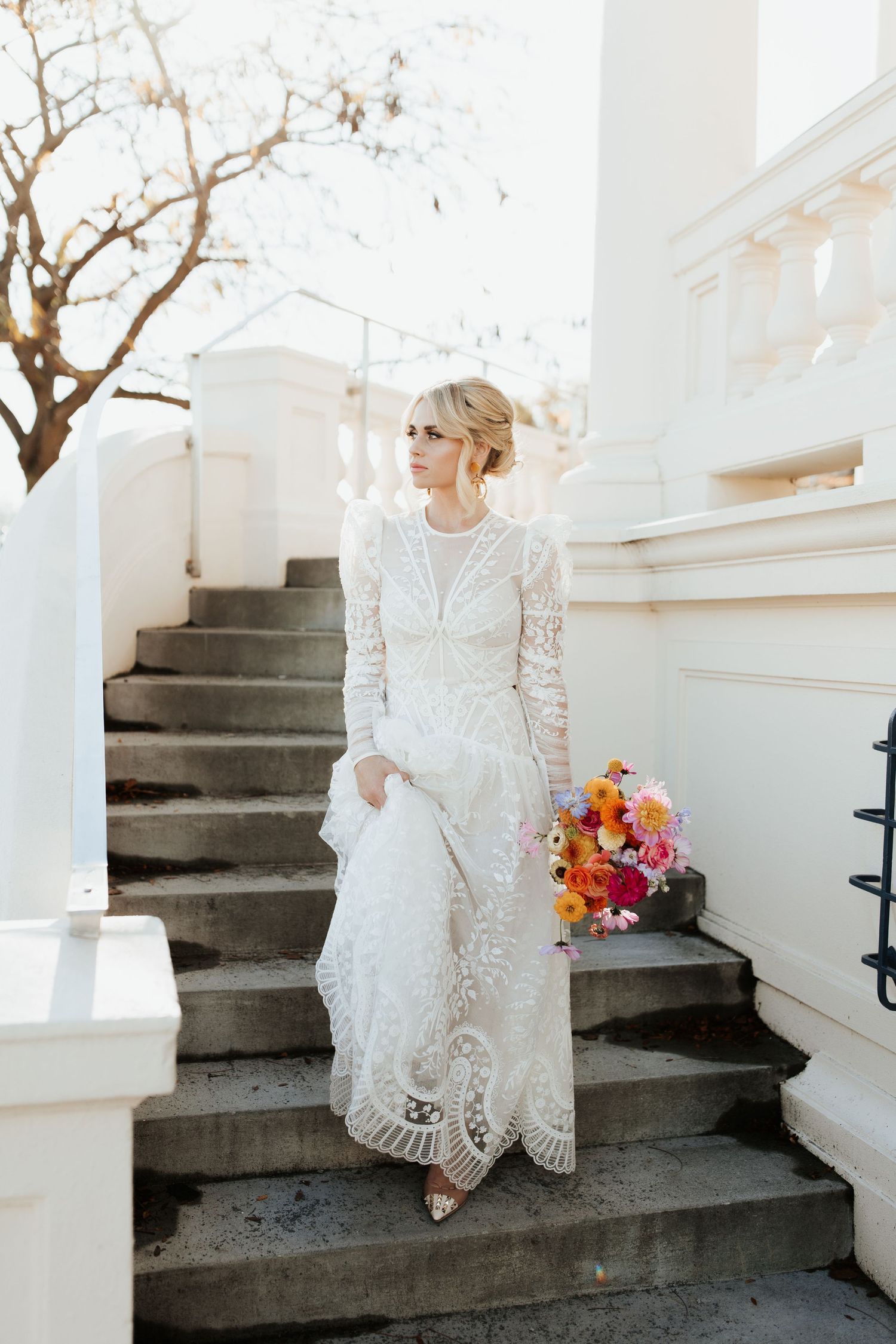 A bridal figure in an elegant white lace dress sits on concrete outdoor stairs, holding a vibrant bouquet.