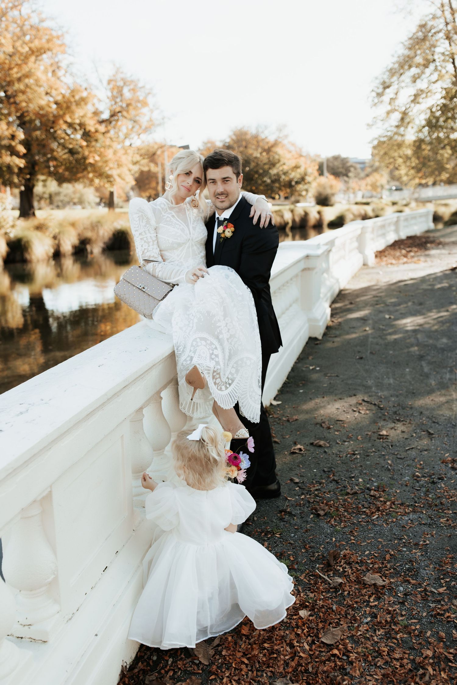 Wedding party in formal attire poses along white bridge railing during autumn sunset.