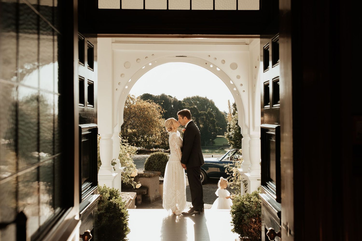 A couple shares a romantic moment under a white stone archway surrounded by greenery and flowers.
