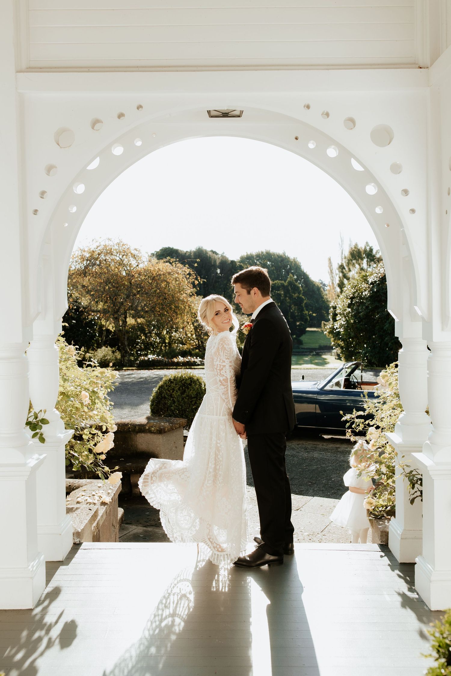A romantic wedding couple stands beneath elegant white archways bathed in golden sunlight.