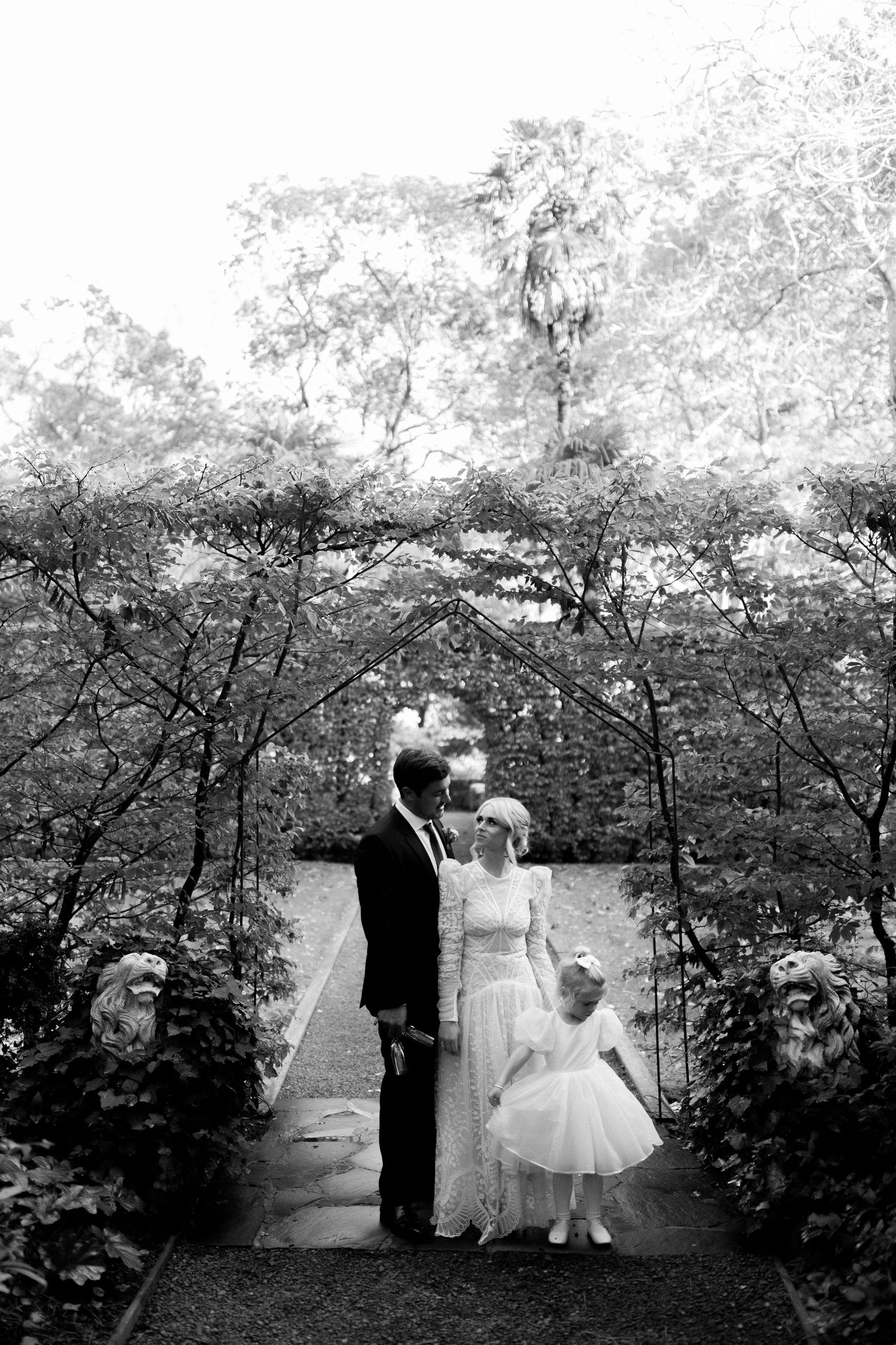 A couple and small child stand on a garden path surrounded by blooming trees and greenery in black and white photography.