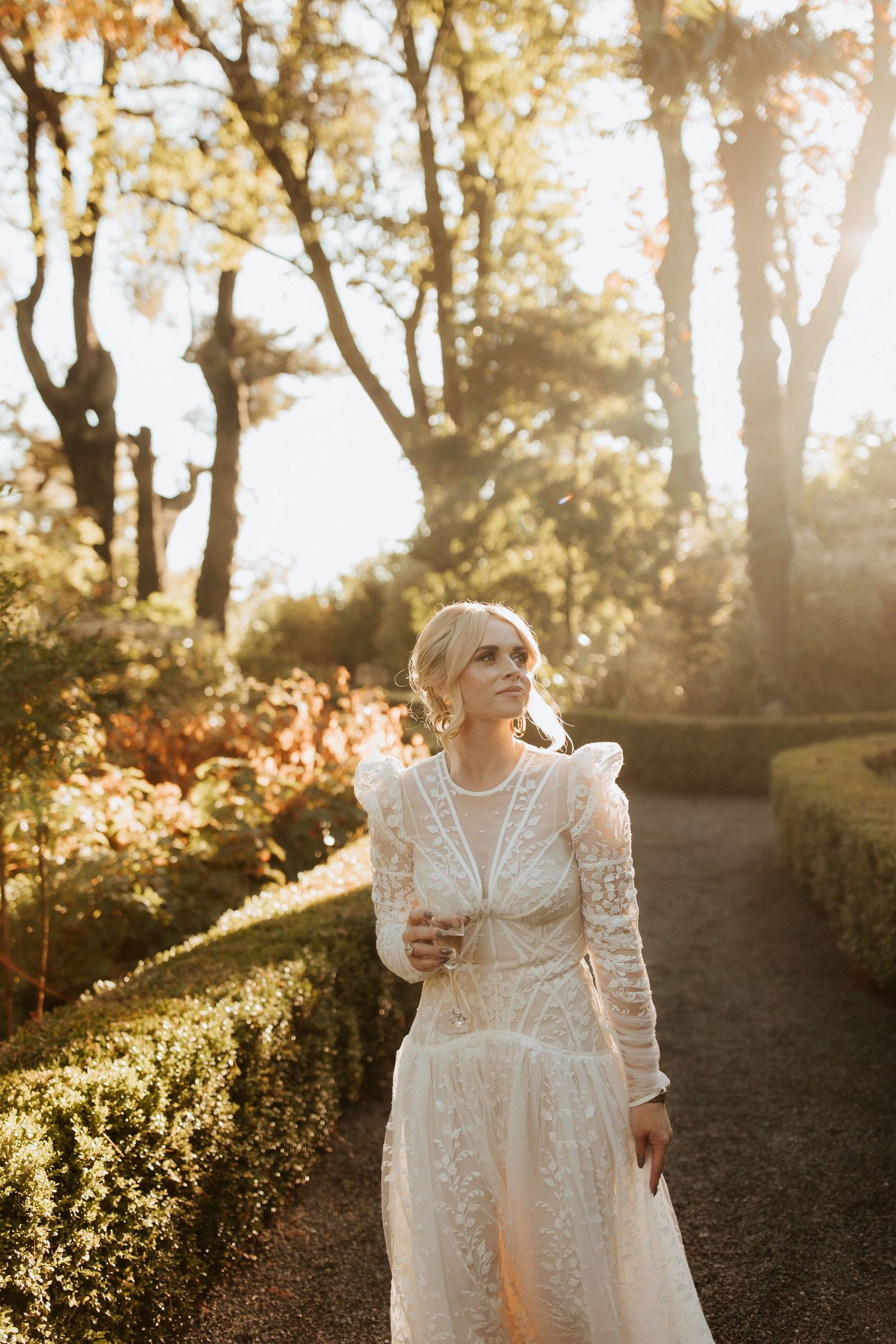 A bride in a long-sleeved lace wedding dress stands on a garden path during golden hour sunset.