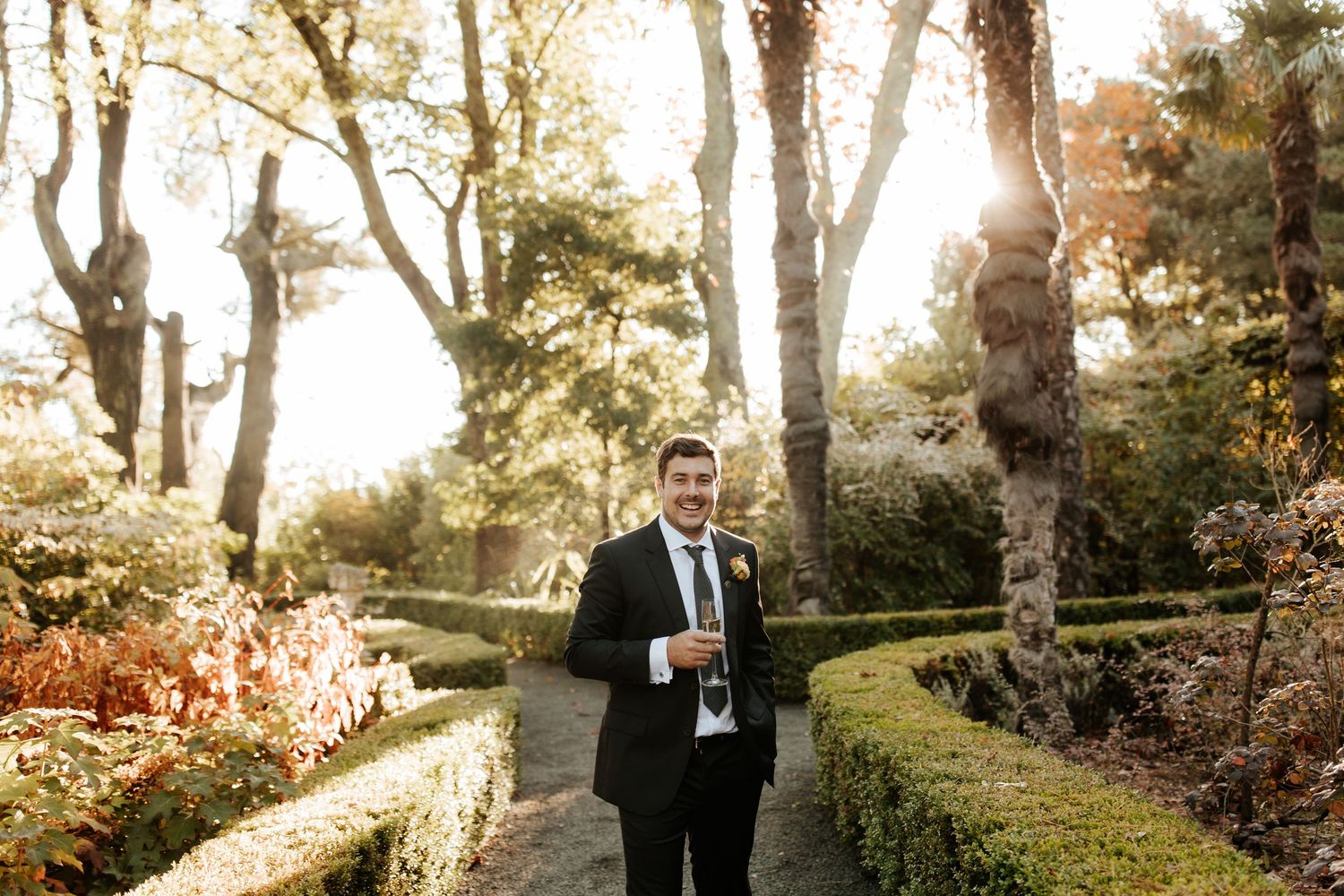 A person in a dark suit holds a drink while standing in a garden path surrounded by autumn trees and hedges.
