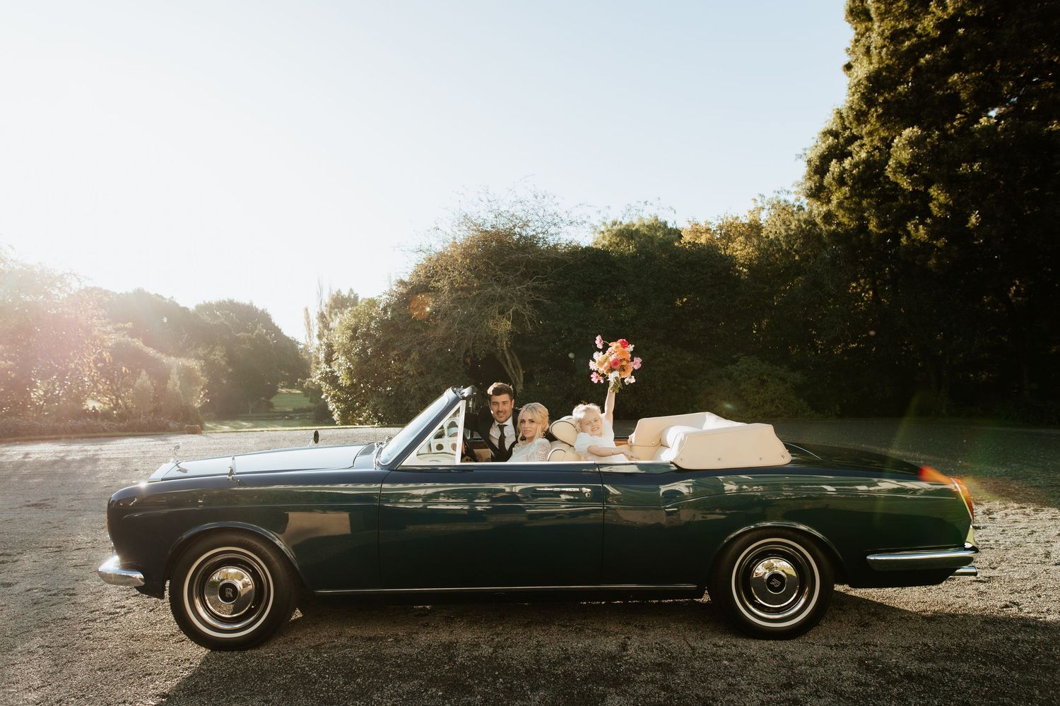 Vintage black convertible car parked on gravel road during golden hour sunset with trees in background.