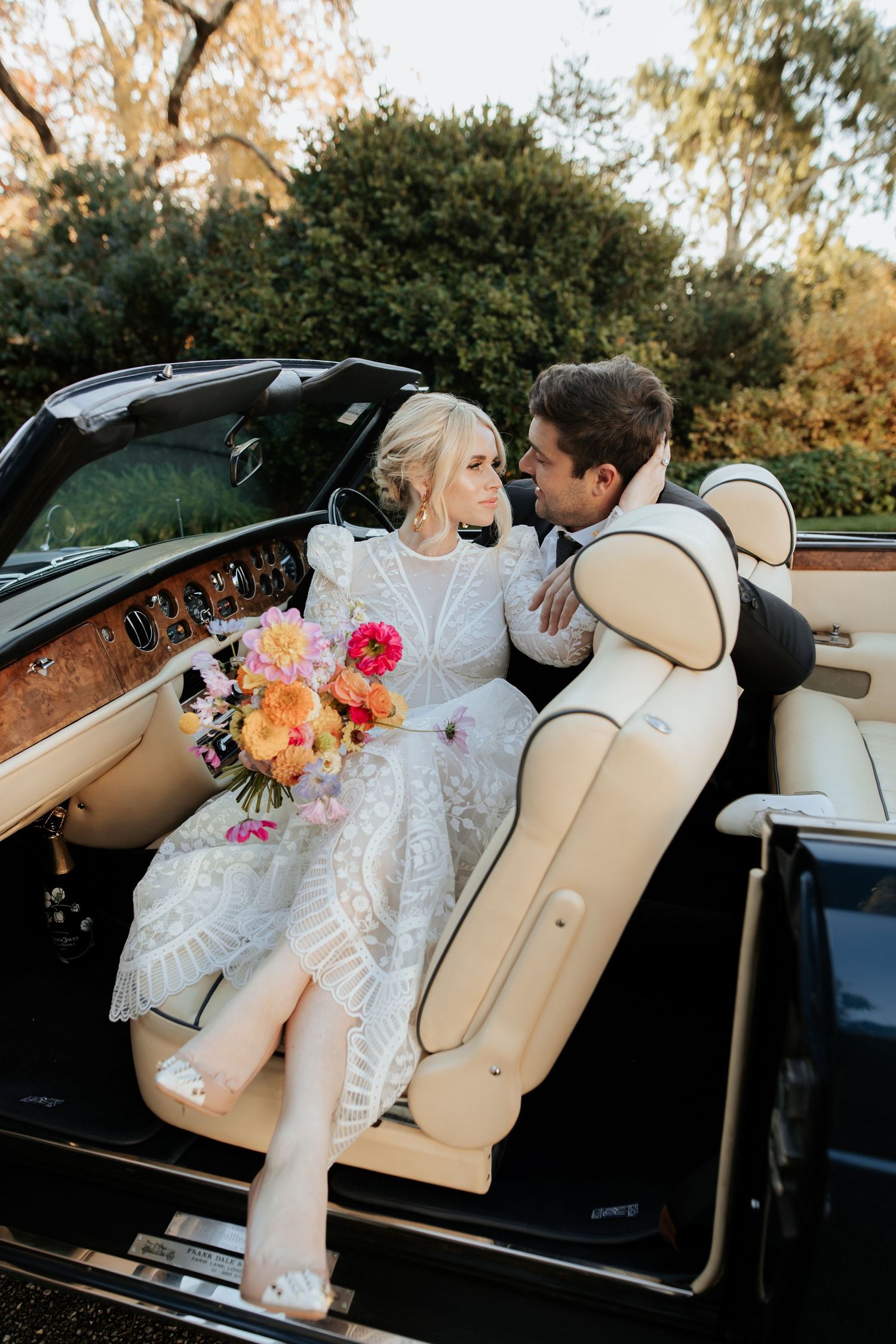 Newlyweds share an intimate moment in a classic convertible car with cream leather seats and colorful wedding bouquet.