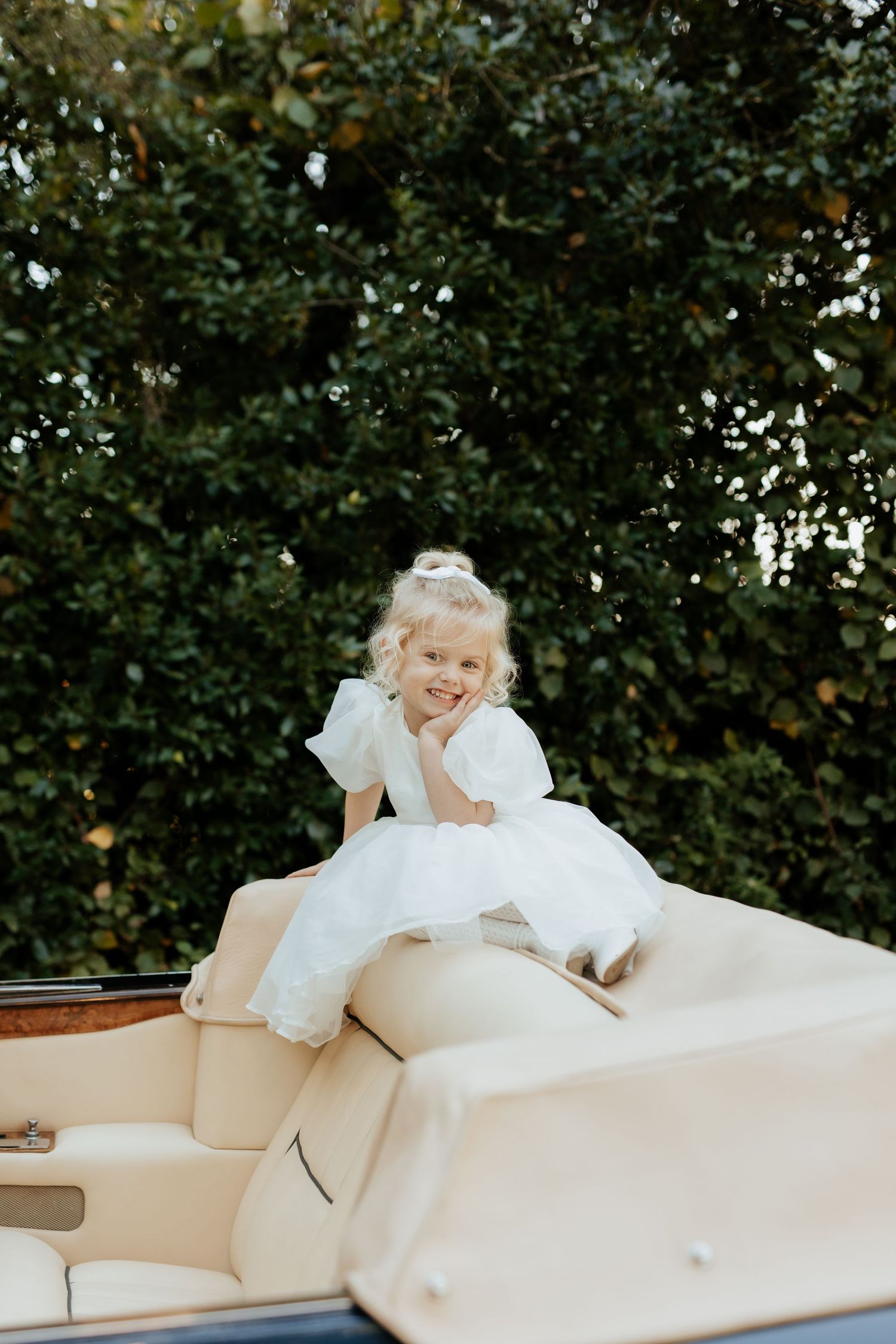 A child in a white dress sits playfully on the back of a cream-colored vintage car in an outdoor setting.