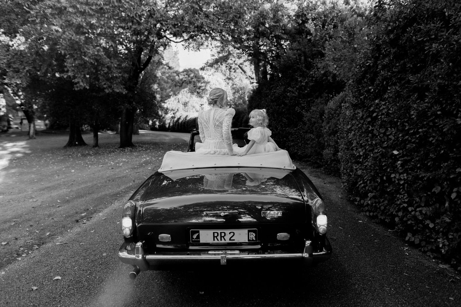 A classic convertible car photographed from behind on a tree-lined street in black and white.