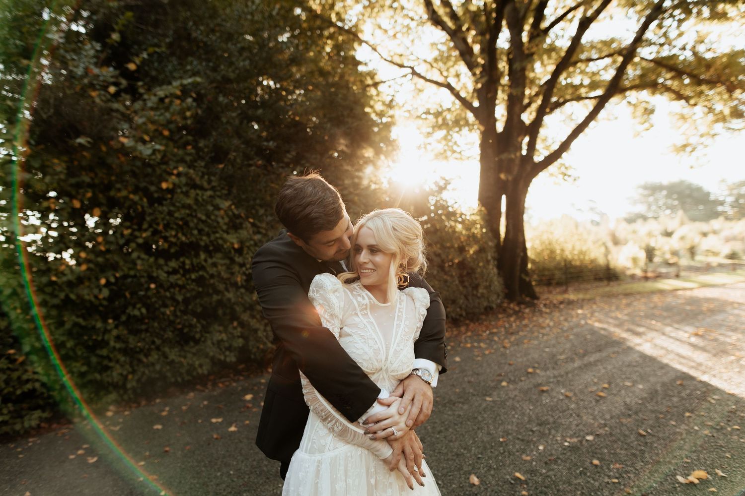 A romantic sunset wedding moment embracing on a tree-lined path with golden sunlight streaming through branches.