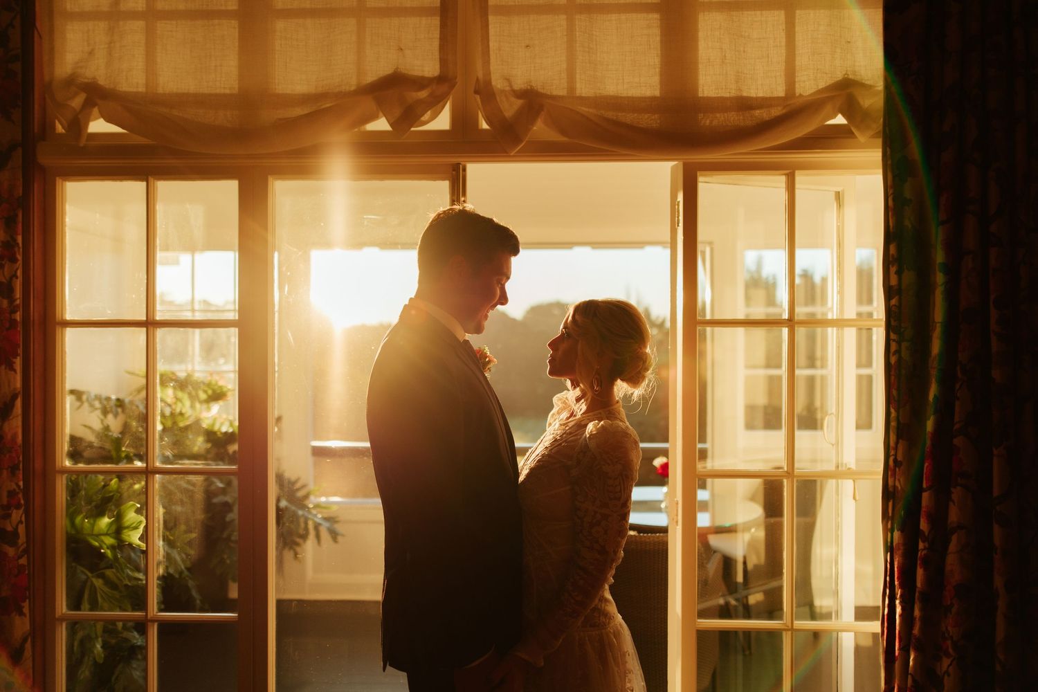 Romantic silhouette of couple embracing by window with warm sunlight filtering through sheer curtains.