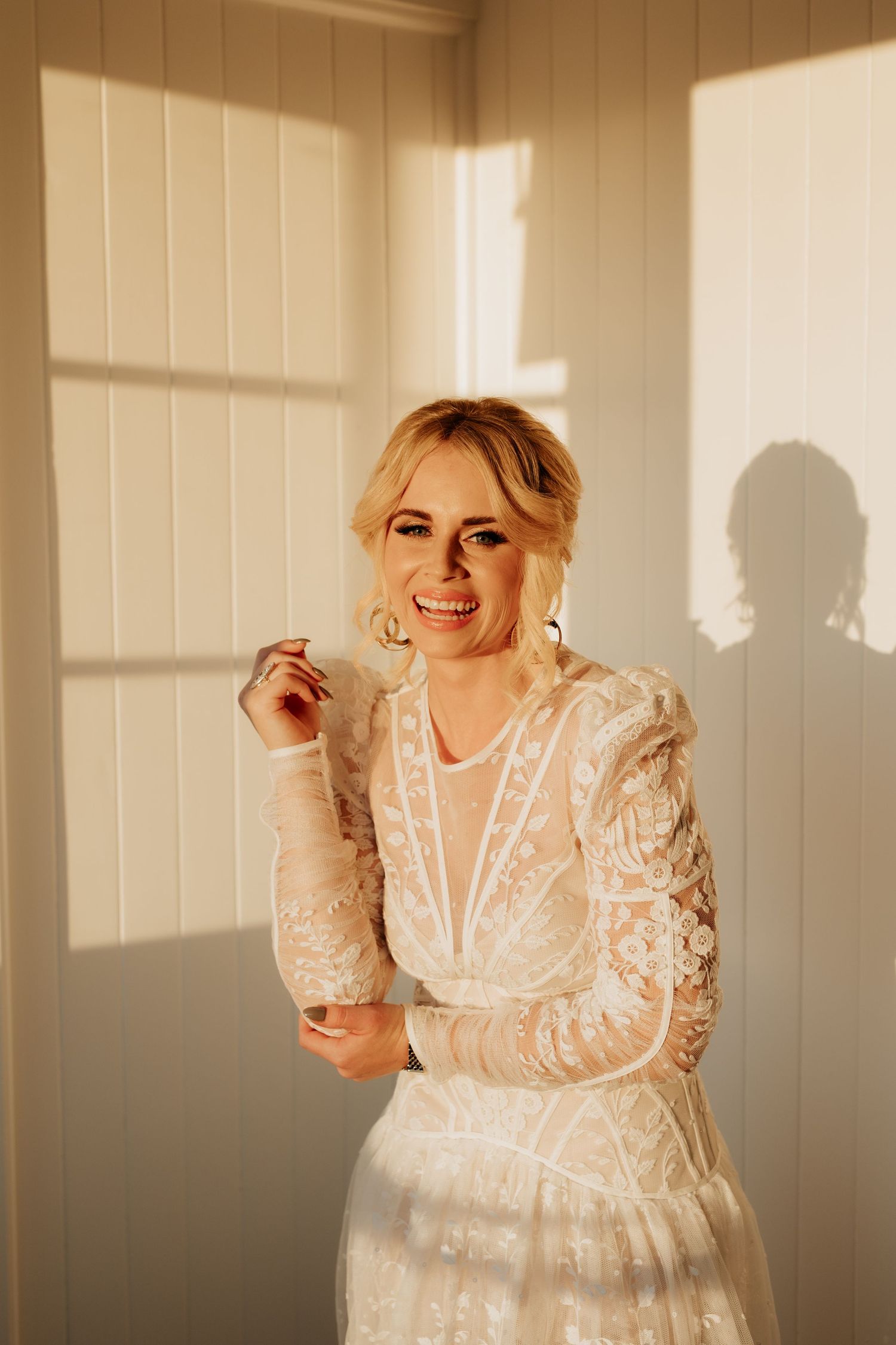A bride in a vintage-inspired lace wedding dress smiles in golden sunlight casting window shadows on a white wall.