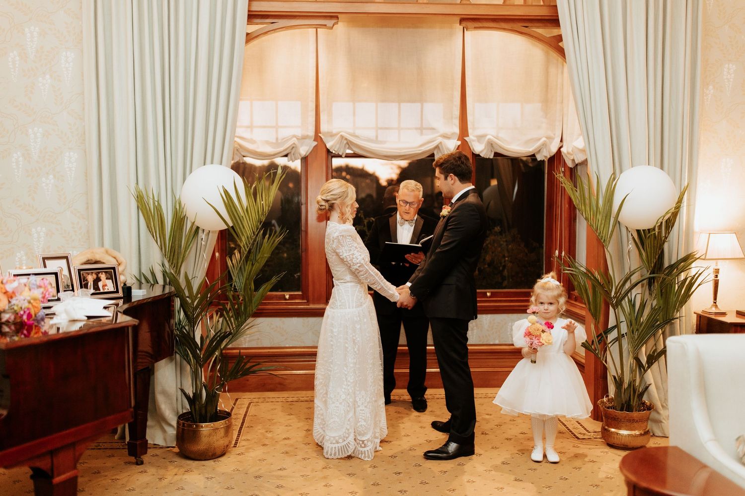 A wedding ceremony takes place in an elegant indoor venue with potted plants and warm lighting.
