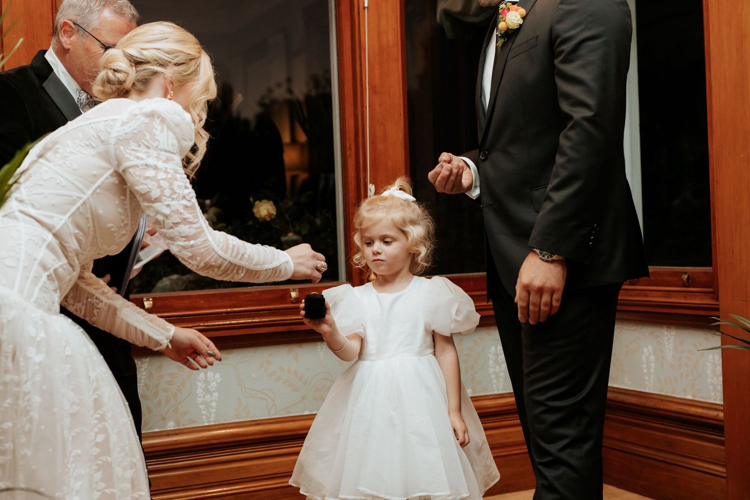 An elegant wedding moment with a little girl in a white dress interacting near a wooden cabinet at a formal event.