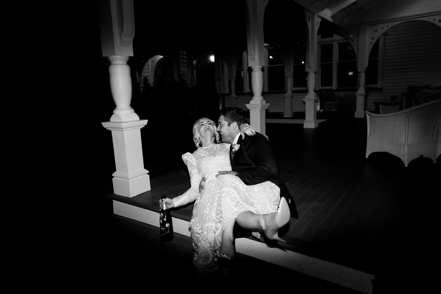 A couple in wedding attire share an intimate moment on a bench in a dark colonnade with illuminated pillars.
