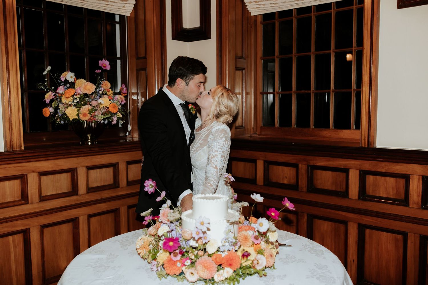 A couple shares an intimate moment behind their elegant wedding cake adorned with fresh colorful flowers in a wood-paneled room.