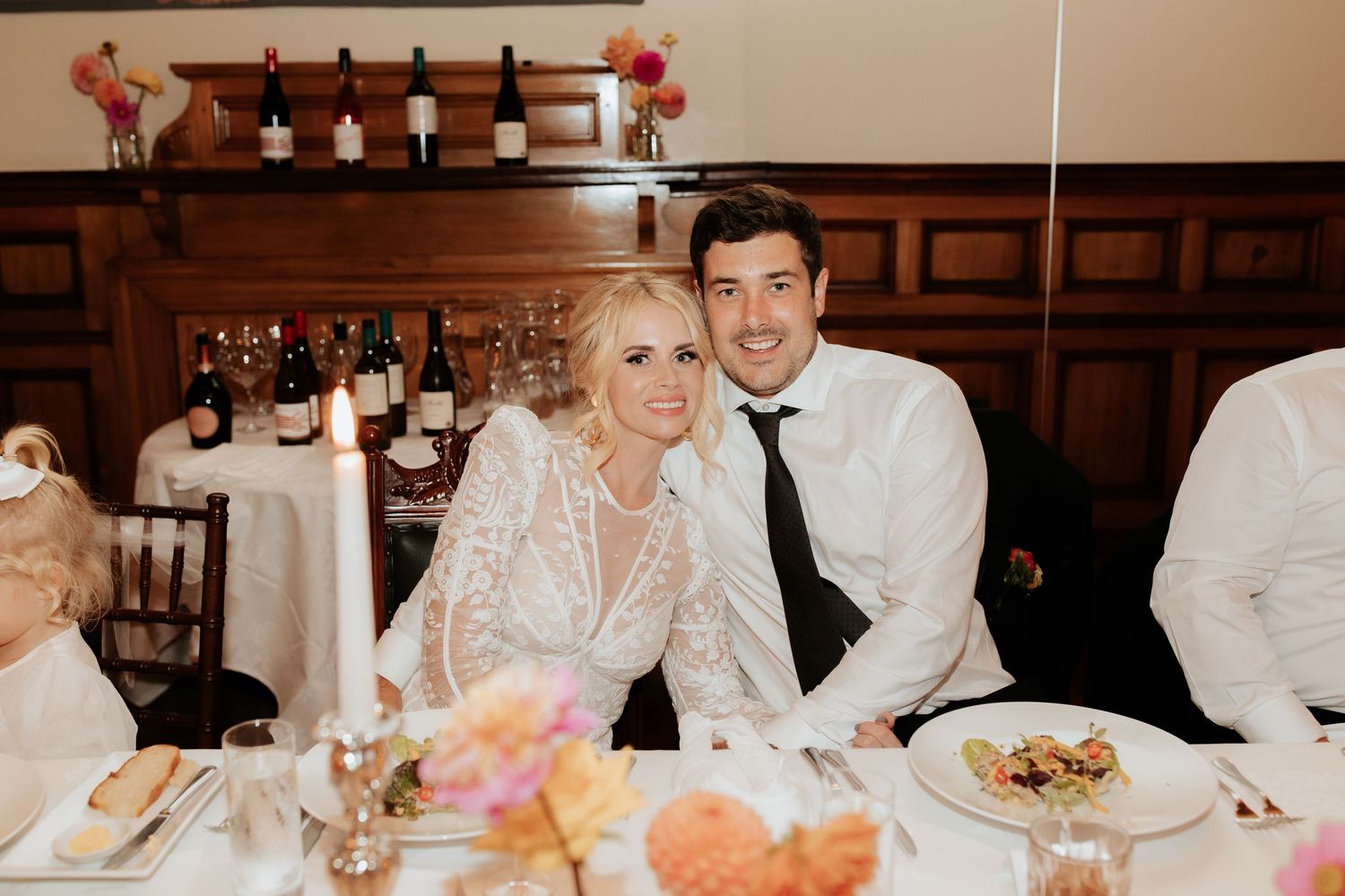A couple sits together at their wedding reception table with drinks and place settings in a wood-paneled dining room.