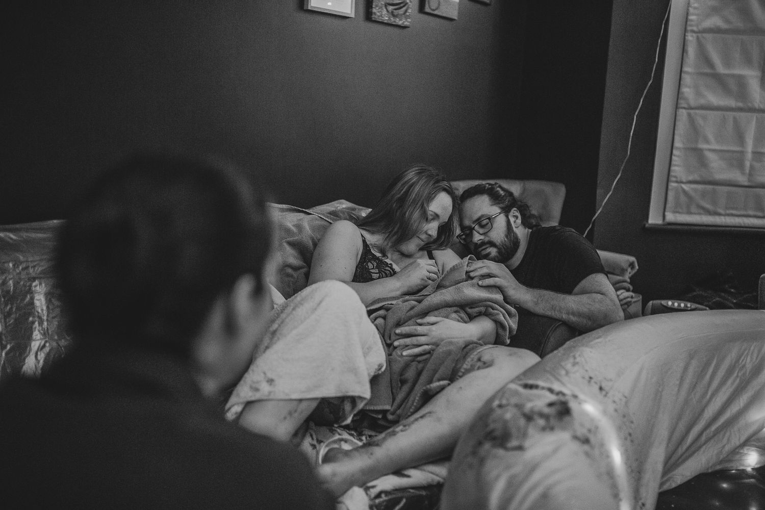 A sweet black and white photo showing a moment of rest with people and a newborn cuddling together on a bed.