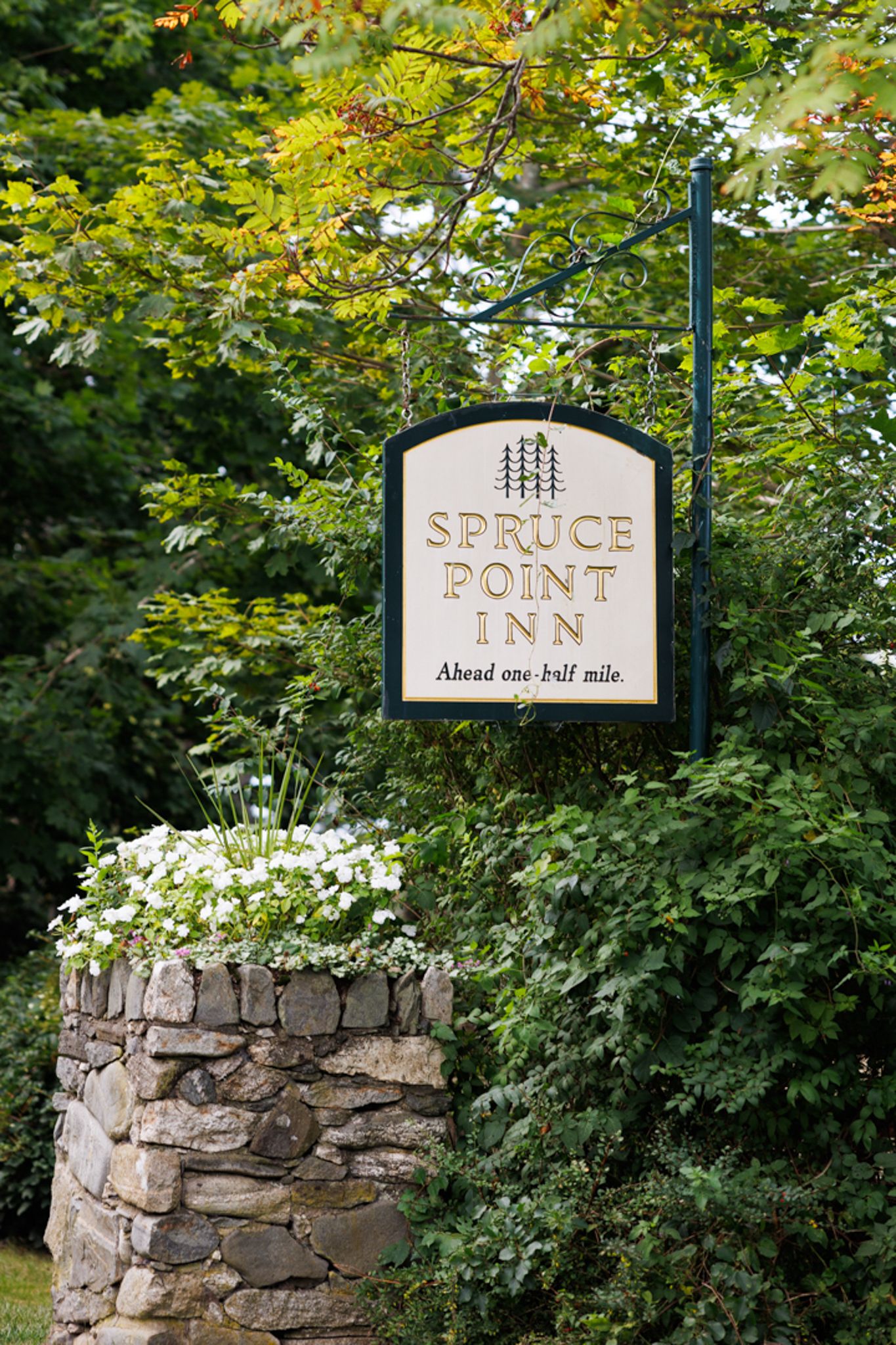 Spruce Point Inn sign mounted on stone pillar surrounded by lush greenery and flowers.