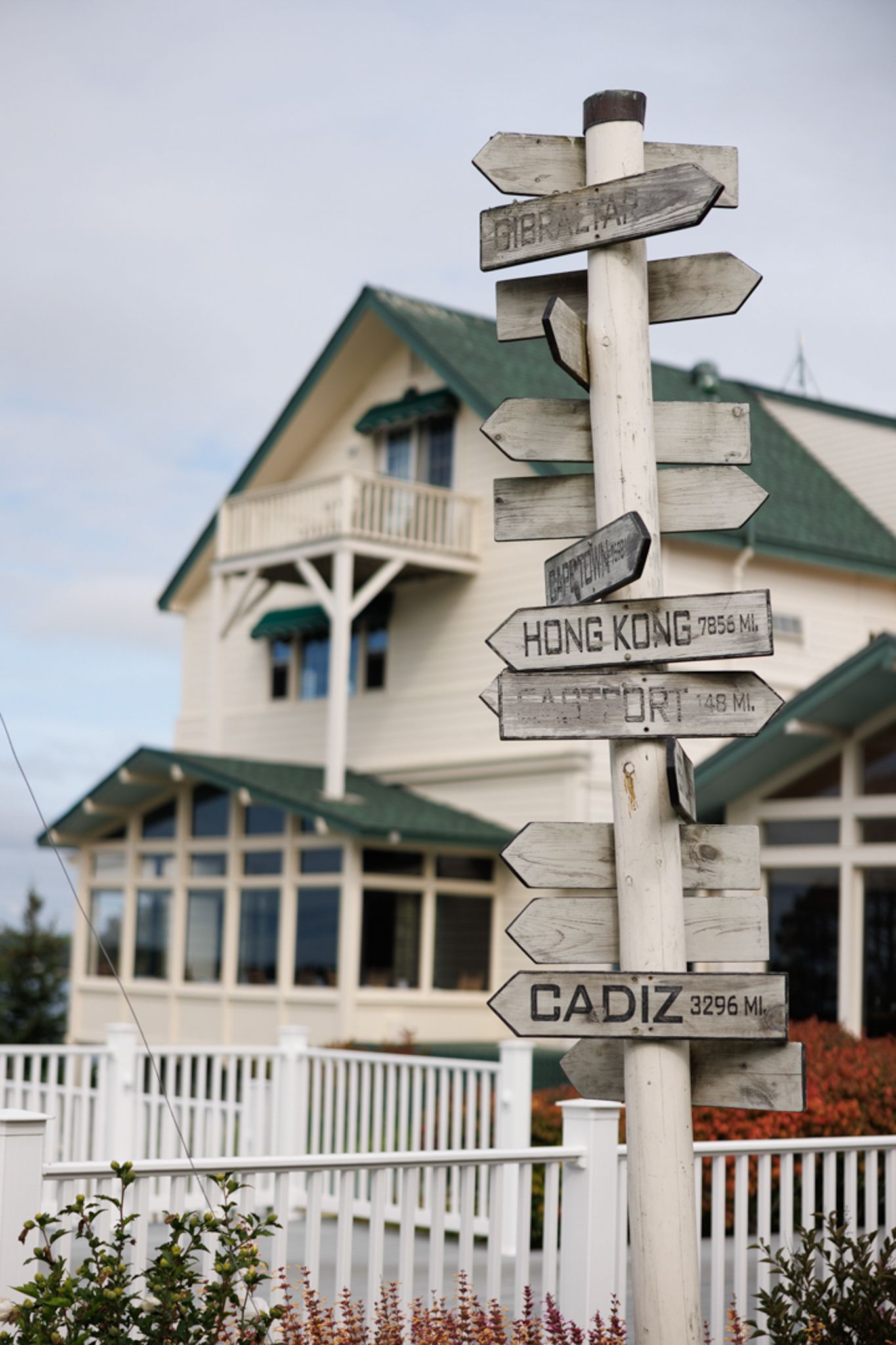 Directional signpost with international destinations in front of a coastal house with white fence.