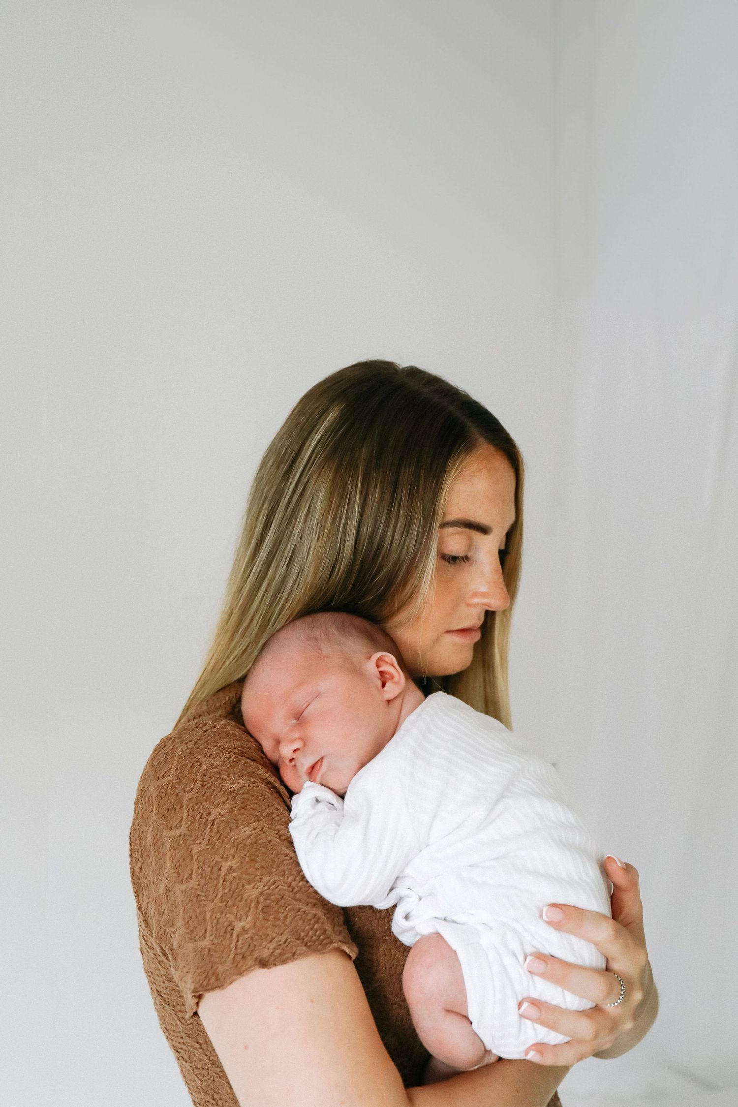 A tender moment shows a baby in white clothing being cradled by mum against a white wall, during newborn shoot based in Nottingham