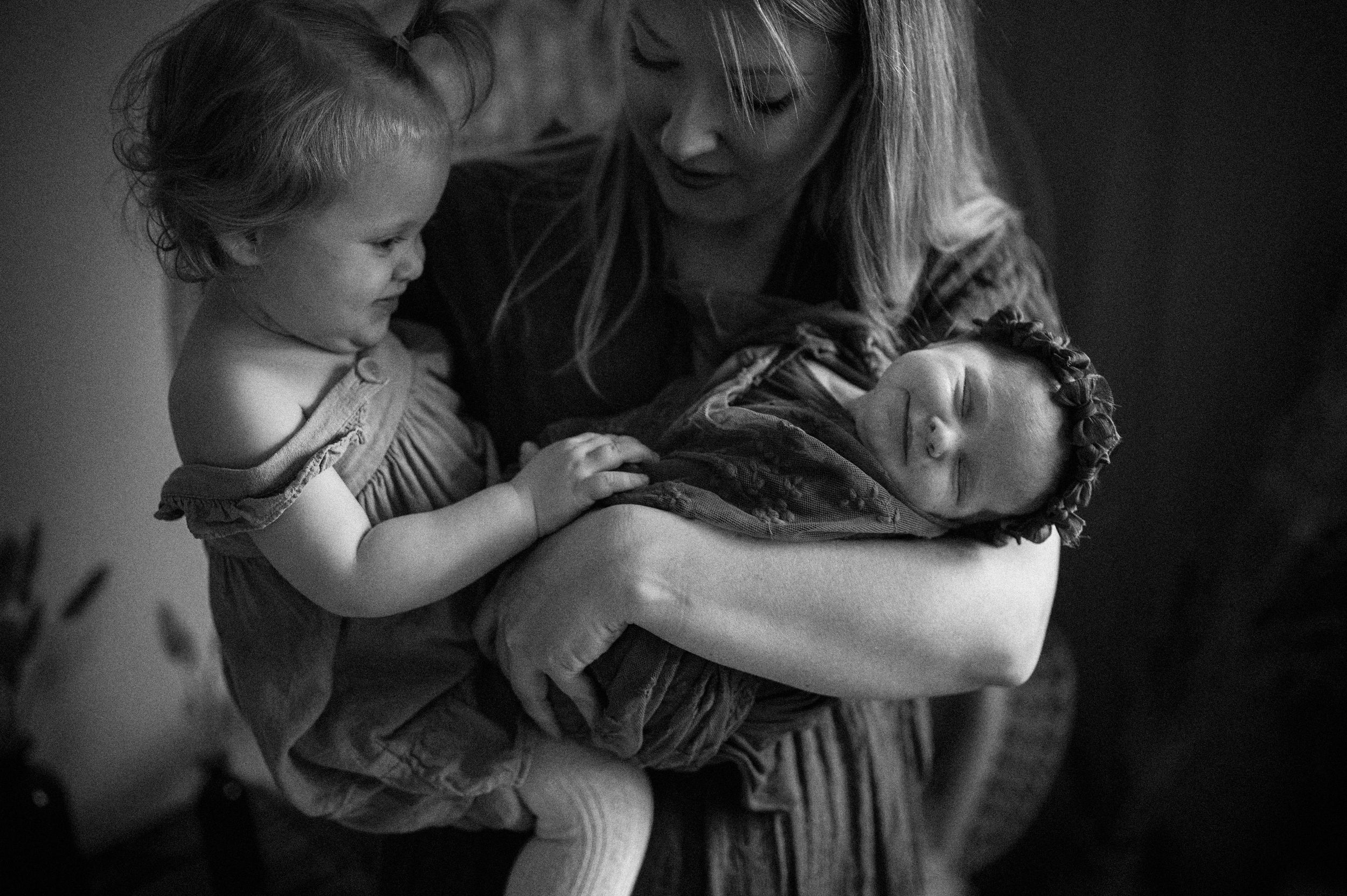 A tender black and white photograph shows a family moment as a child gently touches a newborn in arms.