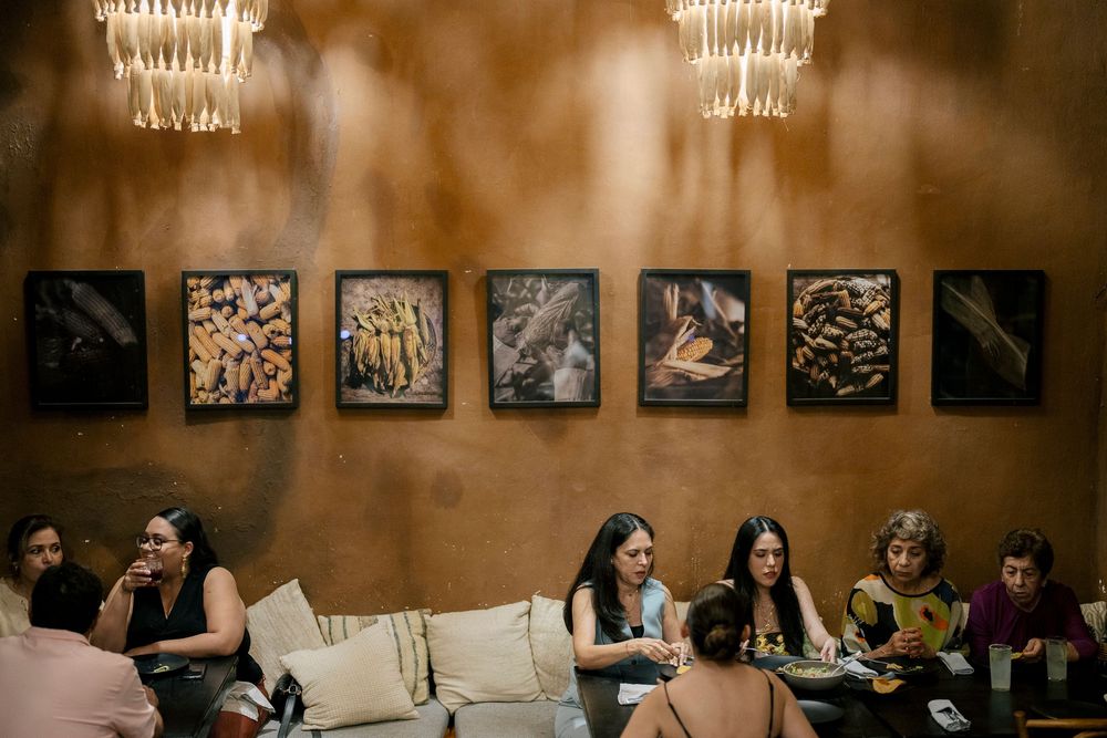 A cozy lounge interior with vintage chandeliers and framed artwork above people gathered on cream-colored seating.