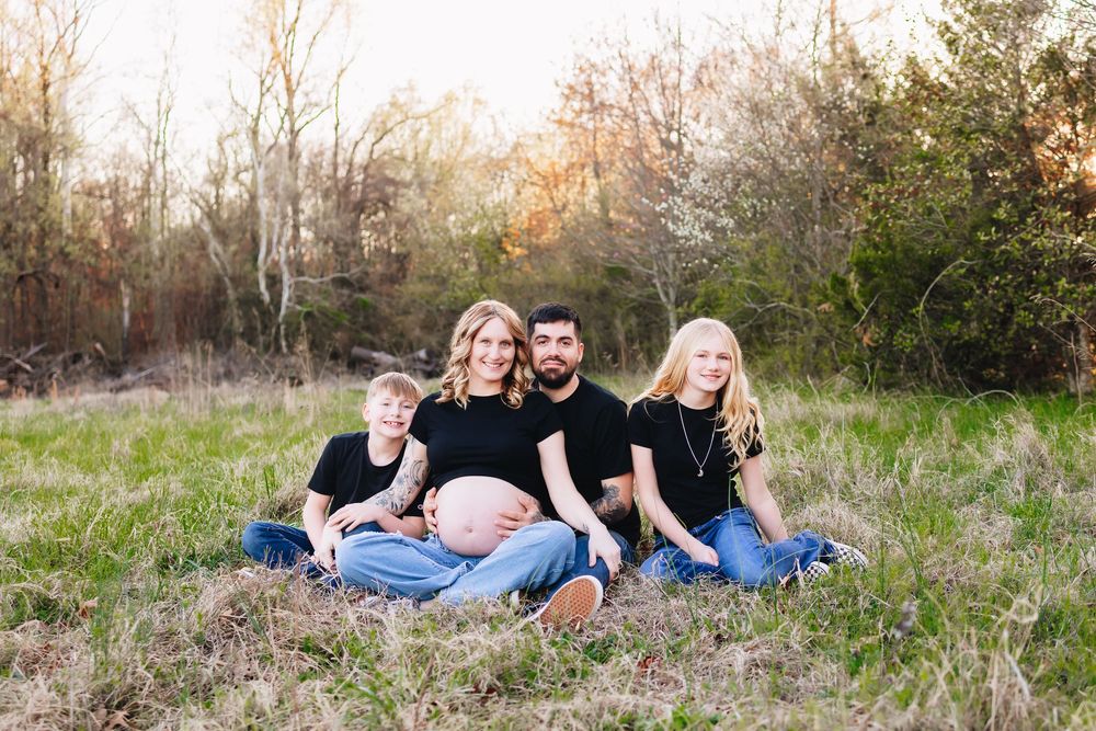 Pregnant woman sits outdoors with family including two children and partner in a sunny green field for maternity photo.