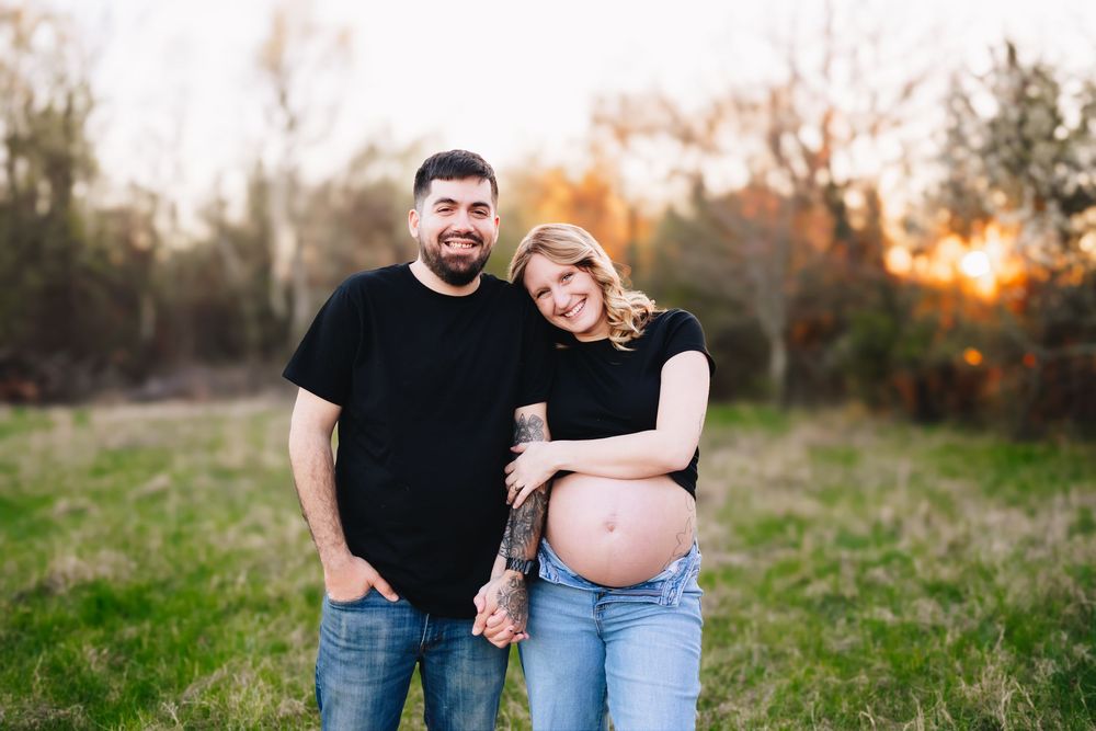 Smiling couple posing outdoors at sunset in a field, woman showing pregnant belly, man in black shirt holding hands.