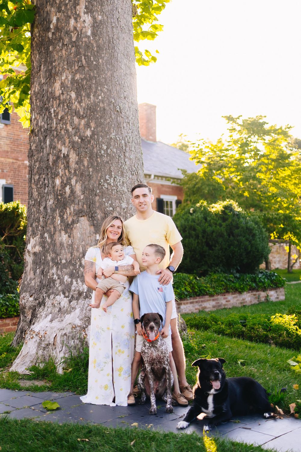 Family portrait with two dogs under large tree near brick building.