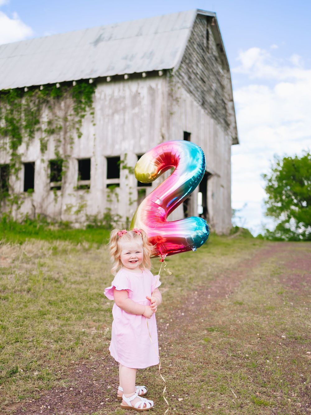 Child holding colorful number 2 balloon in front of weathered white barn.