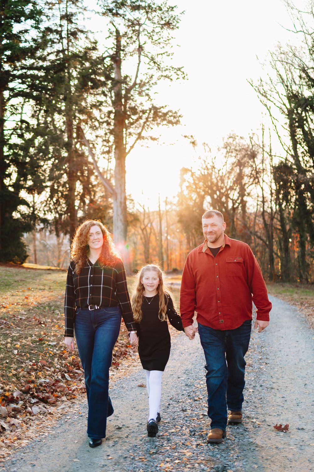 Family of three walking hand-in-hand down a tree-lined path during golden hour in autumn.